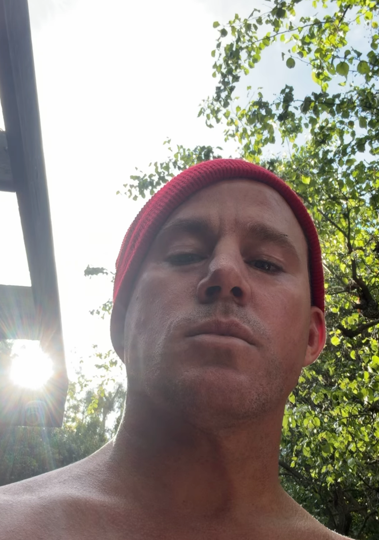 A person outdoors, wearing a knit cap, looking directly at the camera. Sunlight and trees are visible in the background