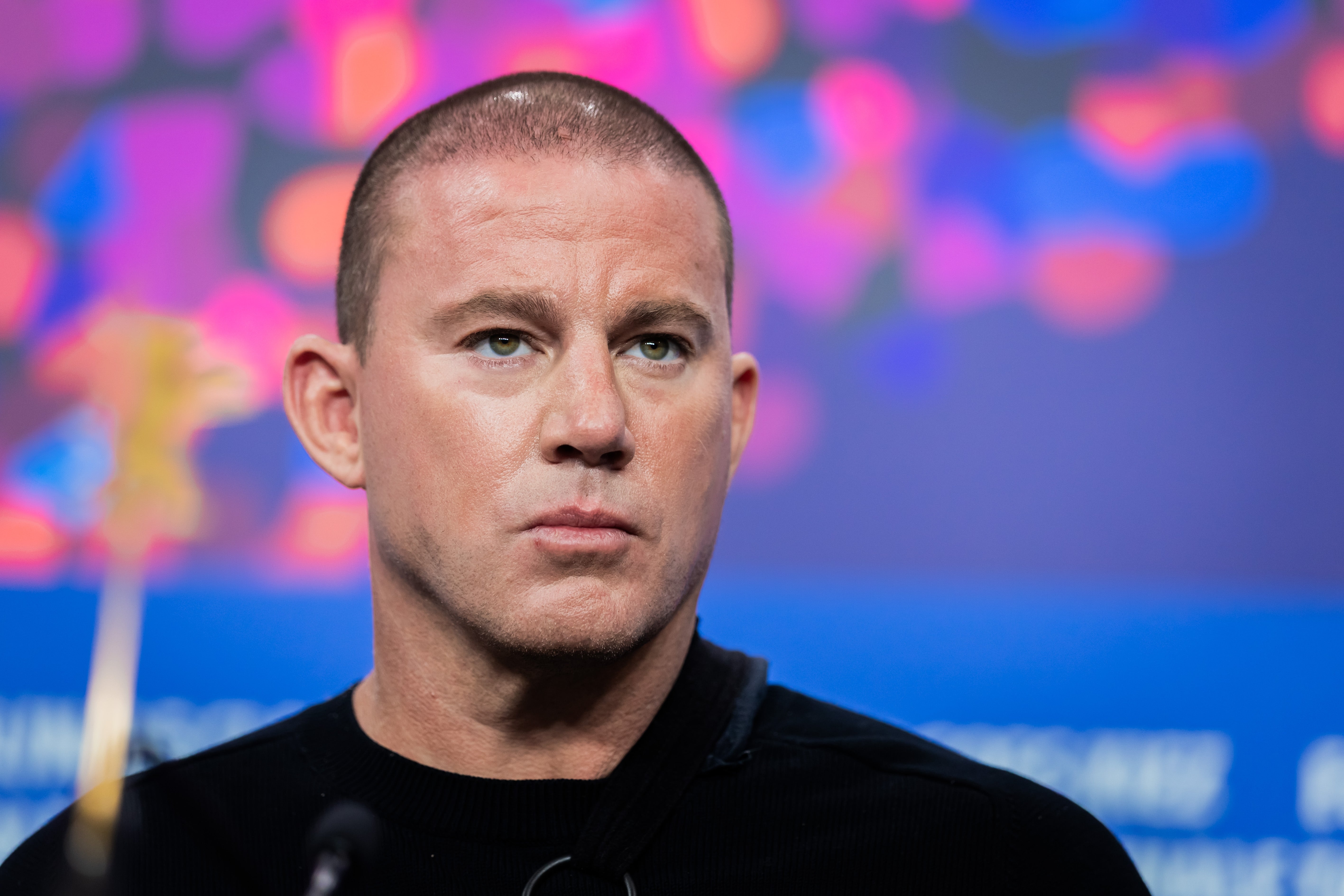 A person with short hair attends a formal event, wearing a classic black top, with a serious expression. Blurred colorful background