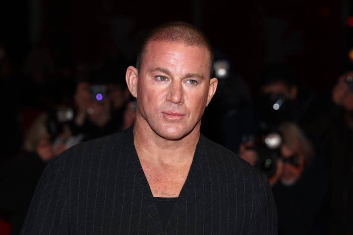 Person on a red carpet wearing a pinstripe suit, photographed by media in the background