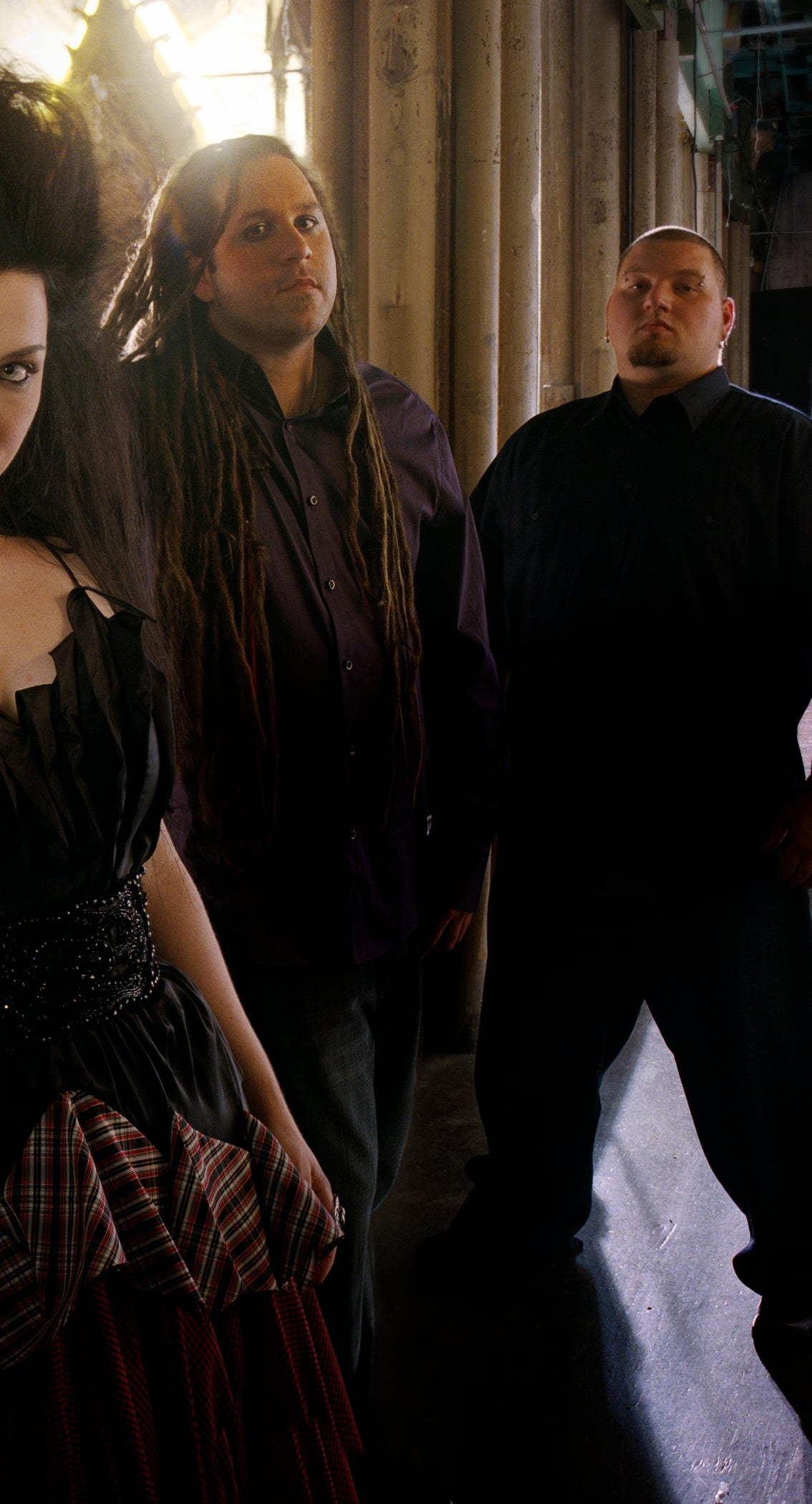 Band poses in an industrial setting. Lead singer wears a tattered dress with layered details. Members stand confidently, reflecting a gothic style