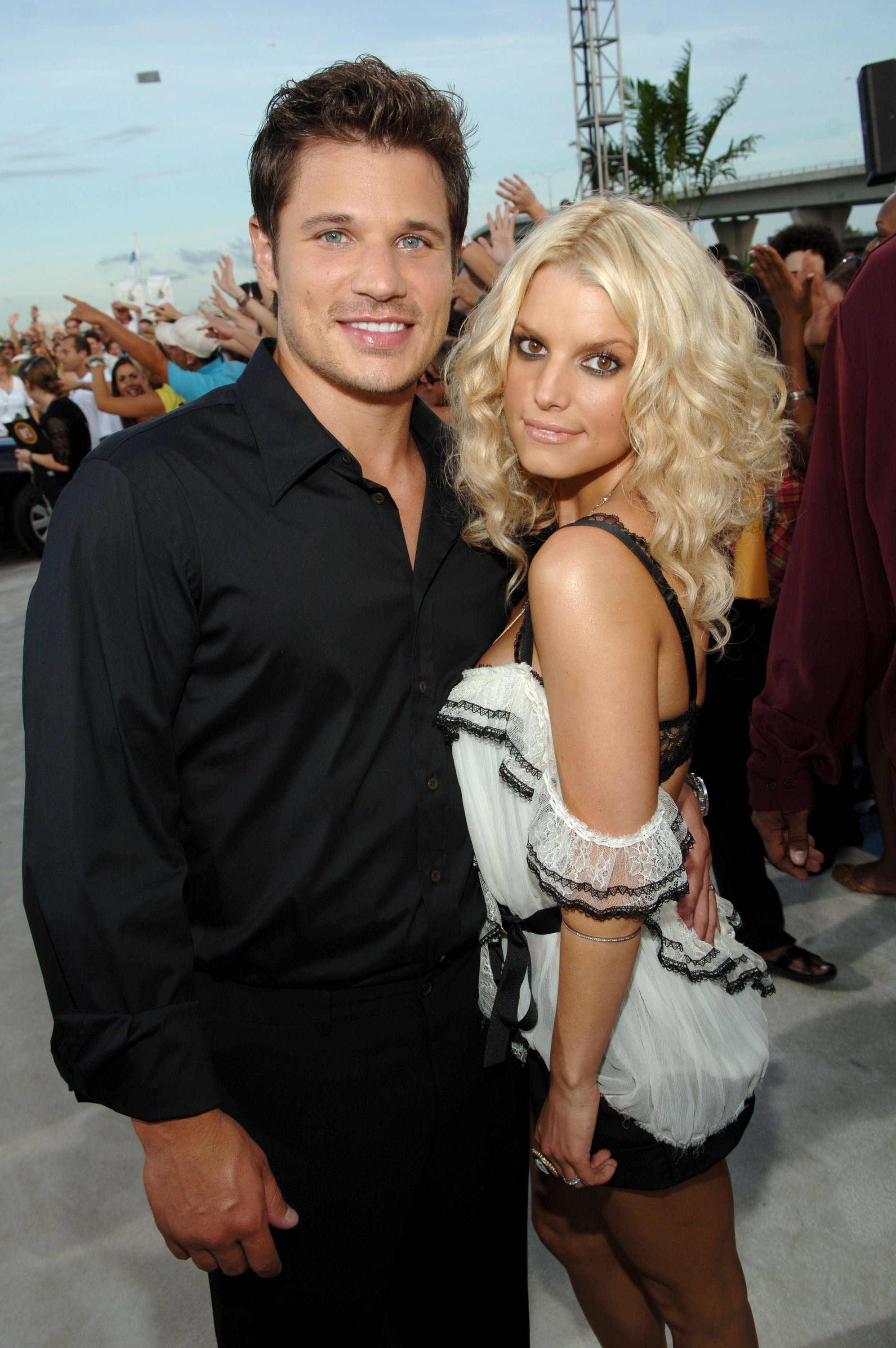 Two people pose together at an event, one in a black shirt and the other in a stylish off-the-shoulder dress. Crowd and palm tree in the background