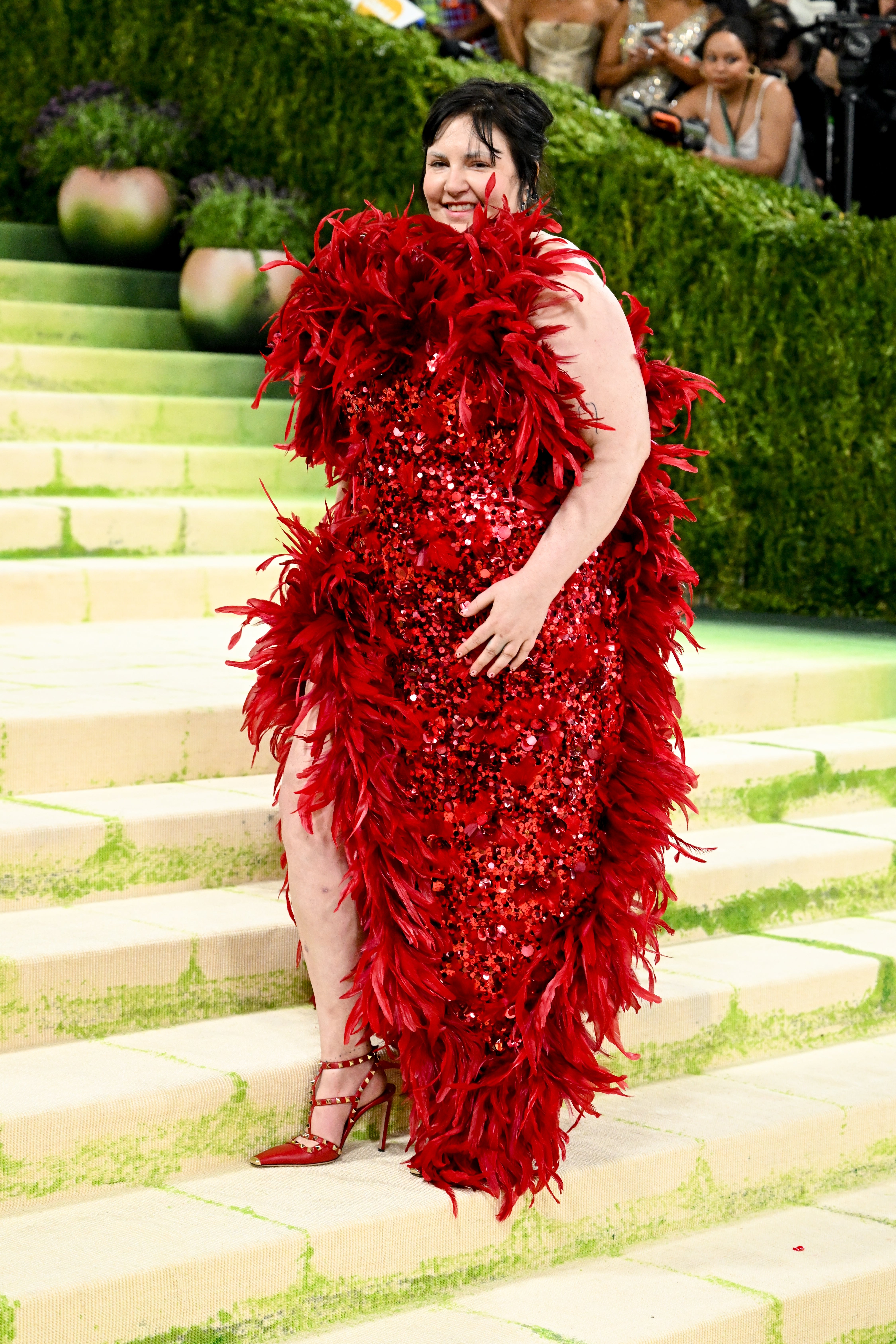 Person in a feathered, sequined dress poses on stairs at a glamorous event, with spectators in the background
