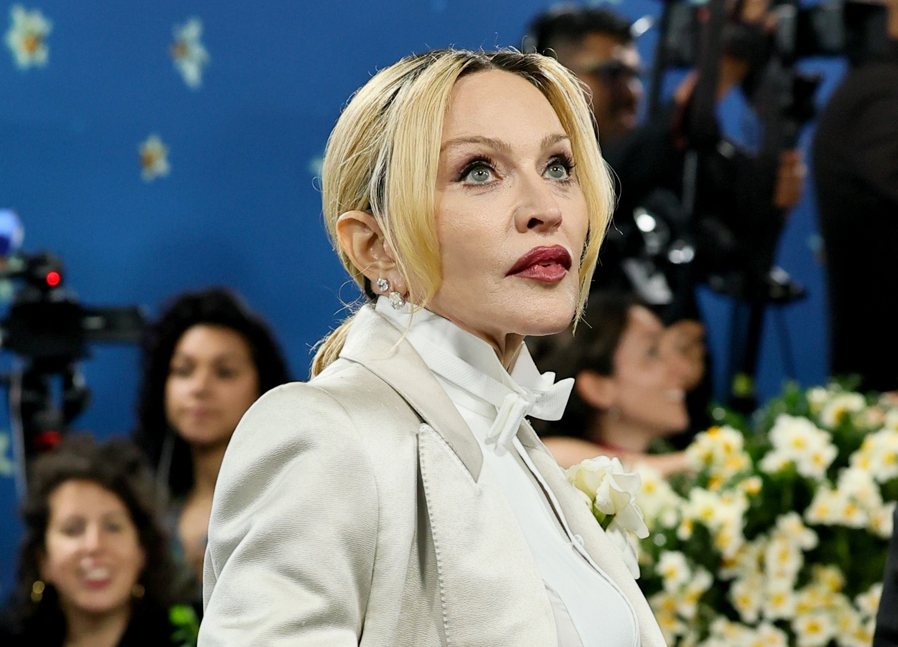 A person with blonde hair is wearing a formal white suit and shirt at a public event. Cameras and people are visible in the background