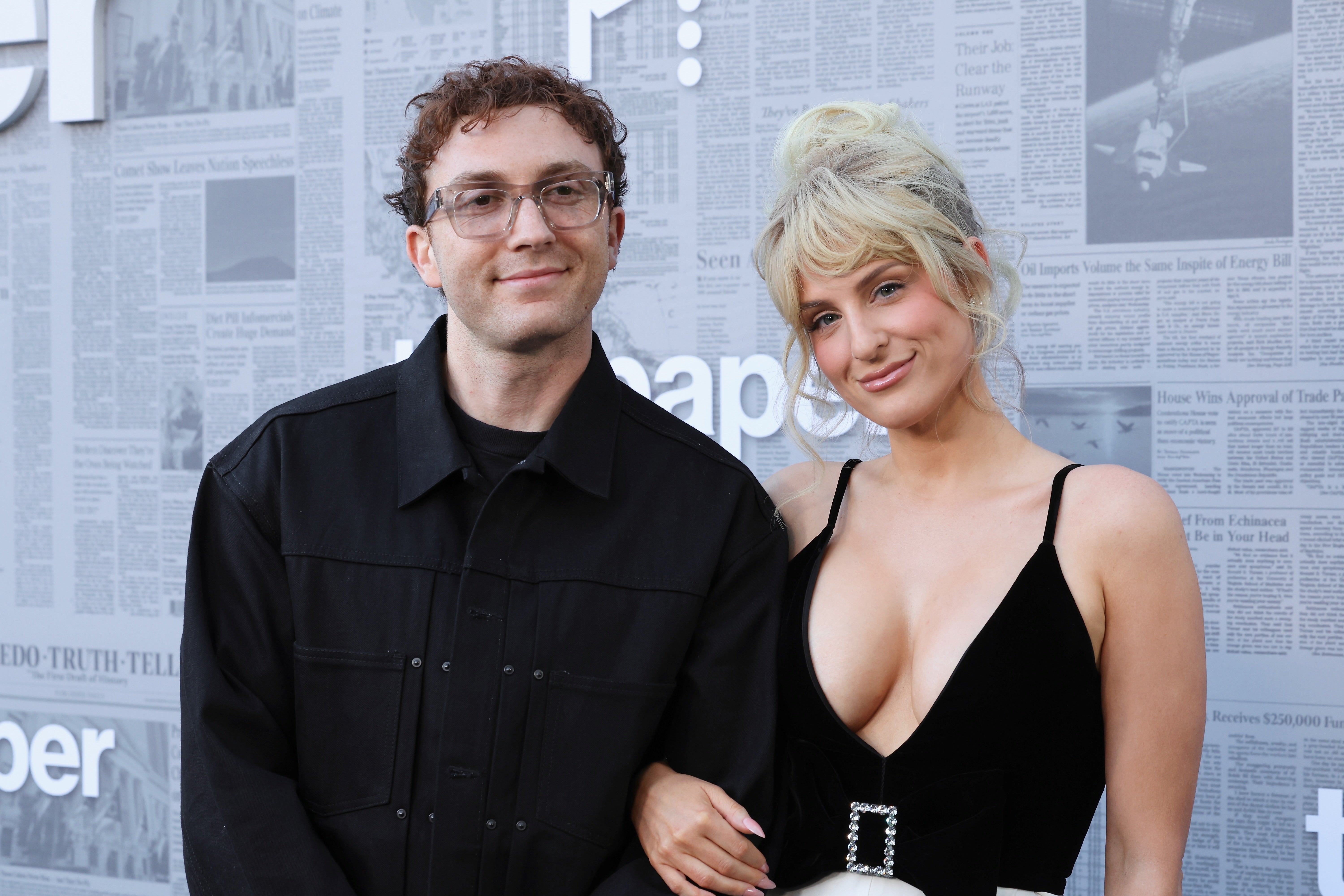 Two people pose together on a red carpet. One is wearing a dark jacket, and the other is wearing a sleeveless dress with a low neckline