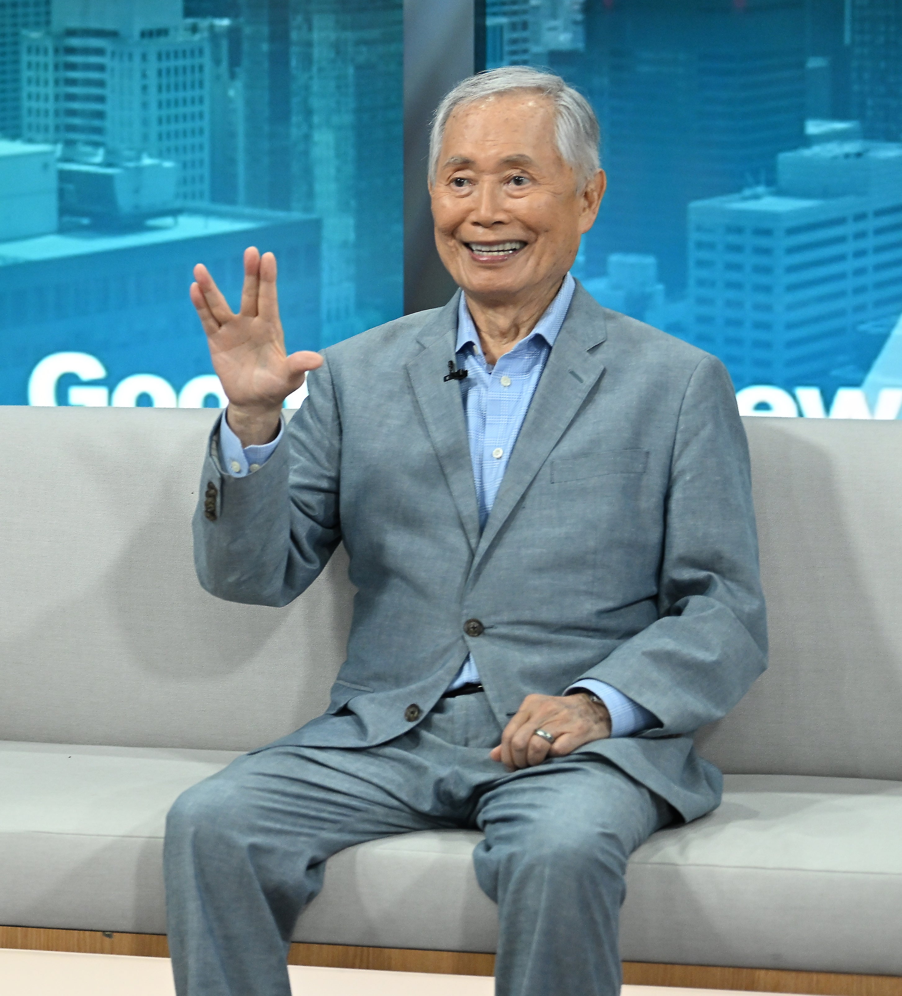 Man in a suit smiling and waving while seated on a talk show set