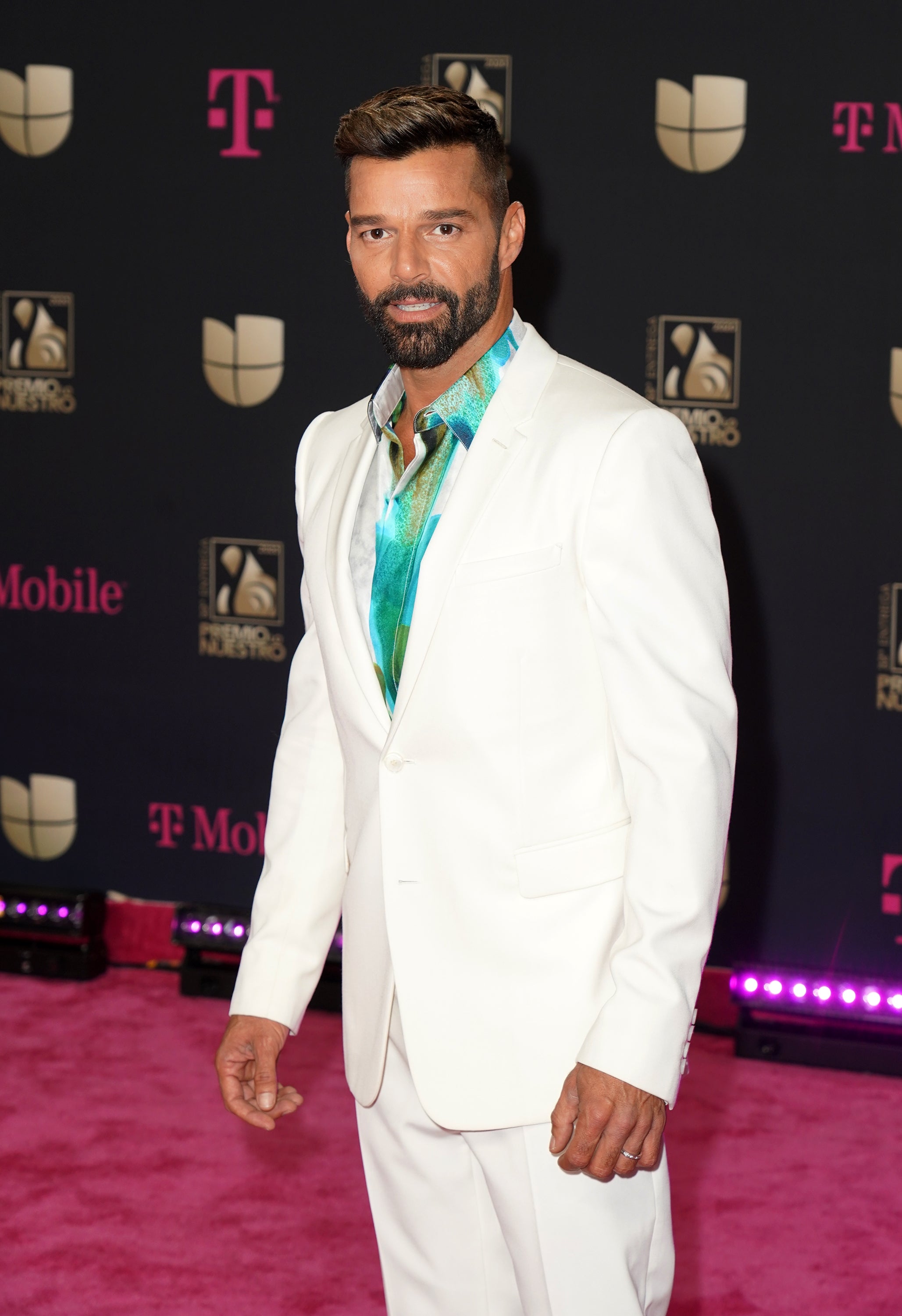 Person wearing a stylish white suit with a bright patterned shirt underneath, attending a formal event with logo backdrop