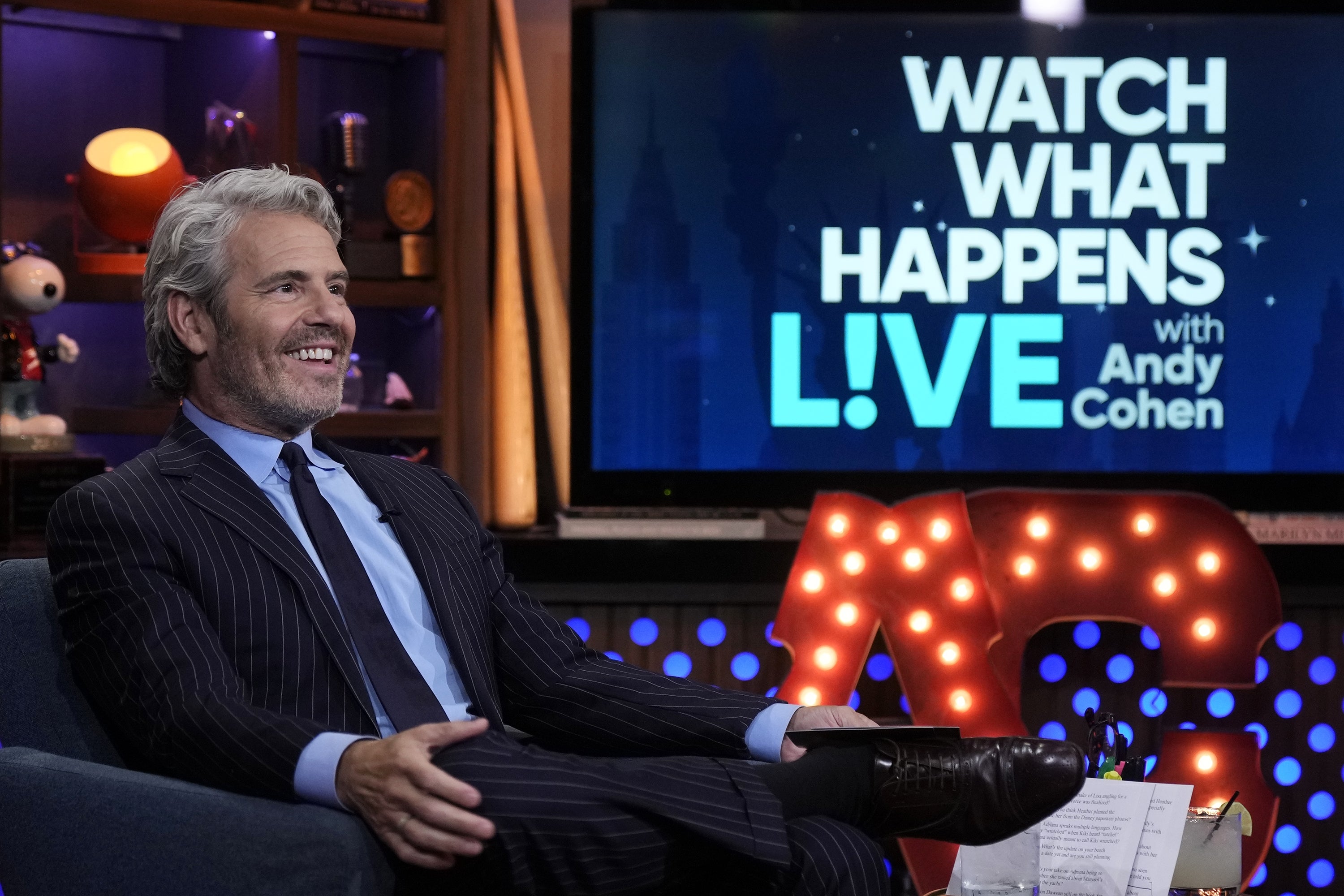 A person in a pinstripe suit sits smiling on a talk show set with a "Watch What Happens Live" sign in the background