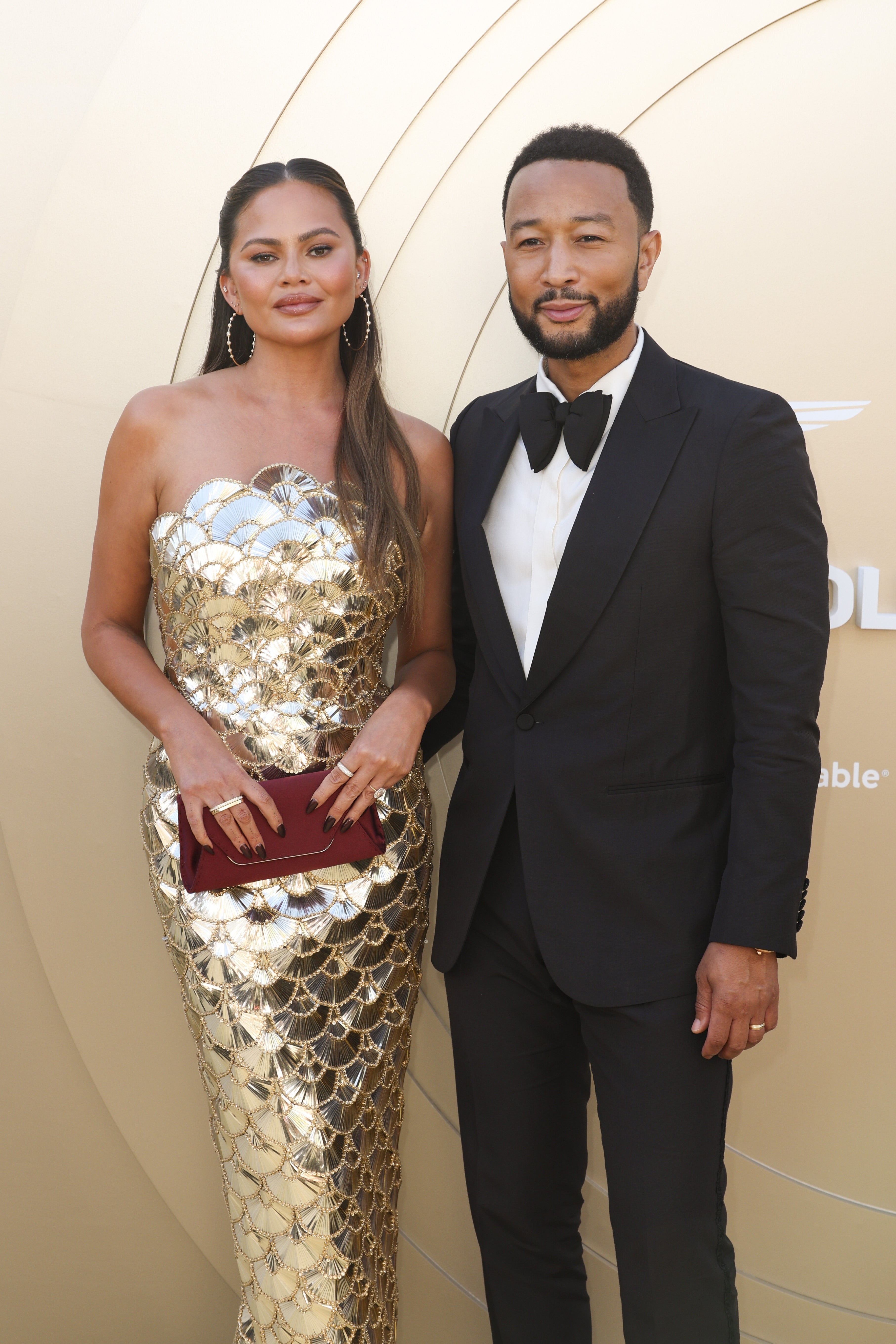 Two people on a red carpet. One wears a strapless, shimmering gown. The other wears a classic tuxedo with a bow tie