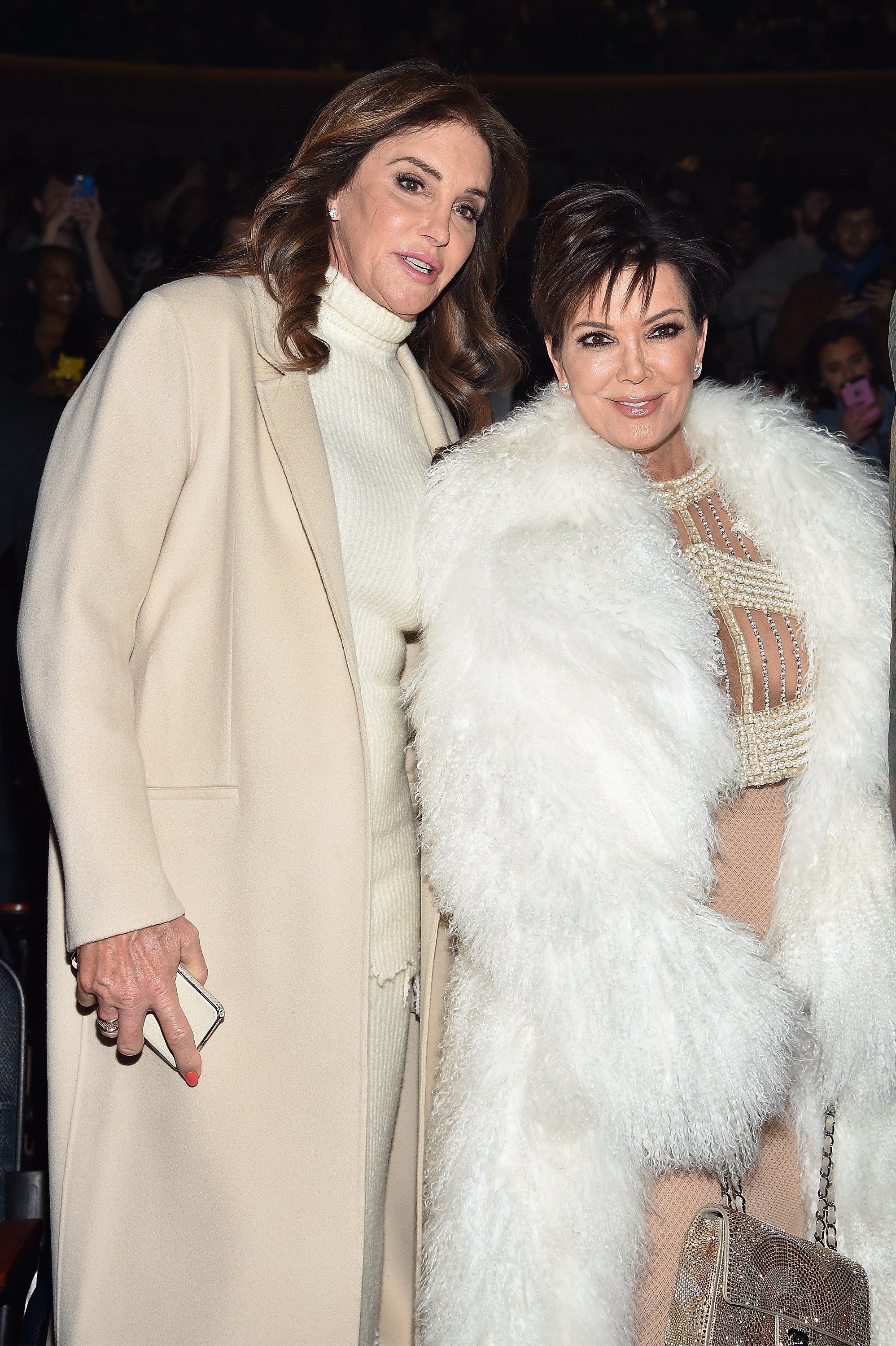 Two individuals on the red carpet; one wears a cream outfit and coat, the other a sheer gown with a white fur coat