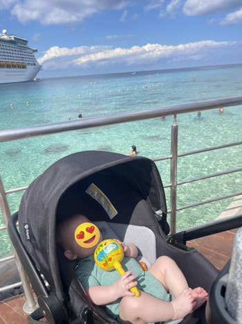 Baby successful stroller holding a toy, adjacent railing overlooking a formation with radical swimming and a cruise vessel successful the distance