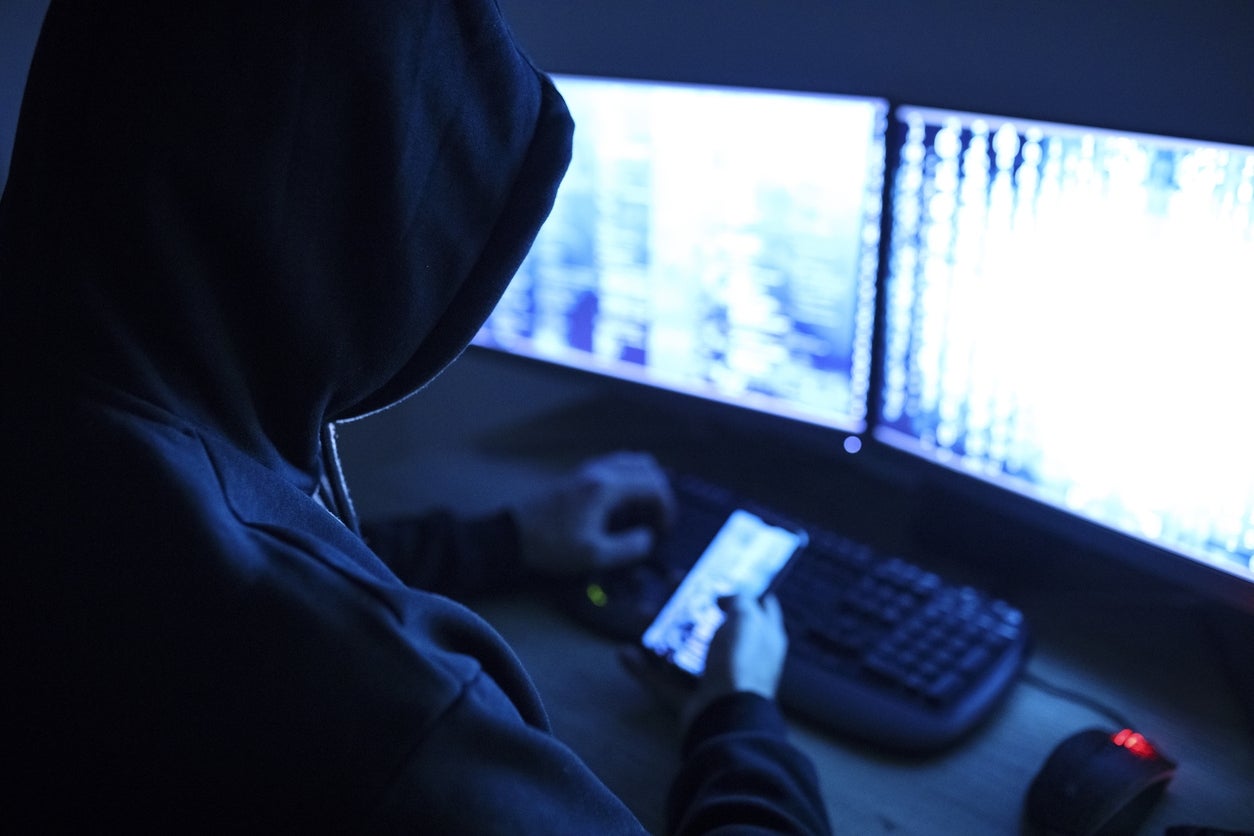 Person i en mørk hettegenser som bruker flere dataskjermer, antyder cyberaktivitet eller hacking
