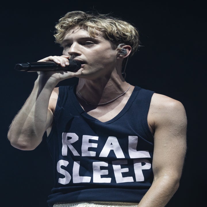 A person is performing on stage, holding a microphone, wearing a sleeveless shirt with "REAL SLEEP" printed on it