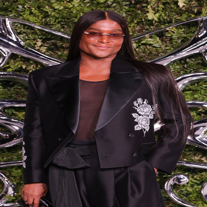 Person in stylish black suit with floral embroidery and sunglasses, standing in front of an abstract silver and greenery backdrop