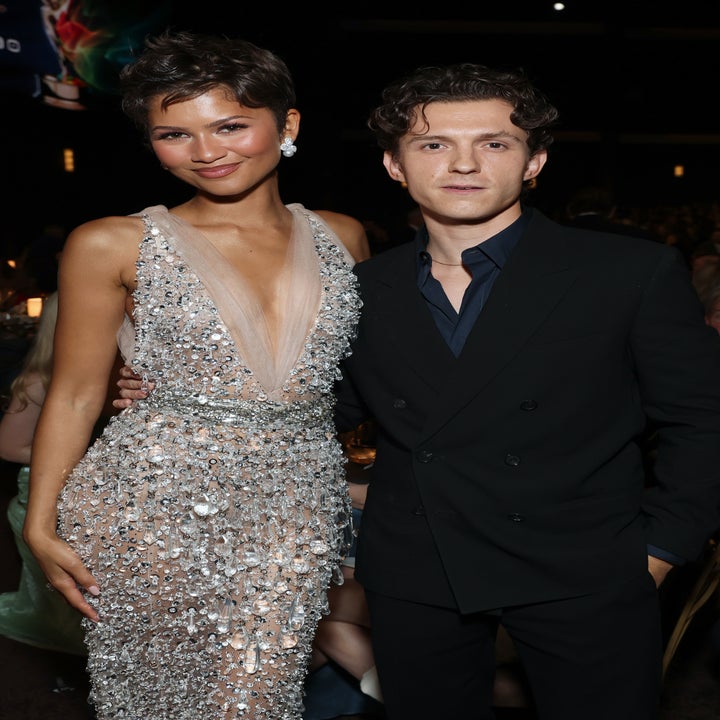 Two individuals pose at an event. One wears a sparkling, sleeveless gown, while the other wears a dark suit. Both are smiling