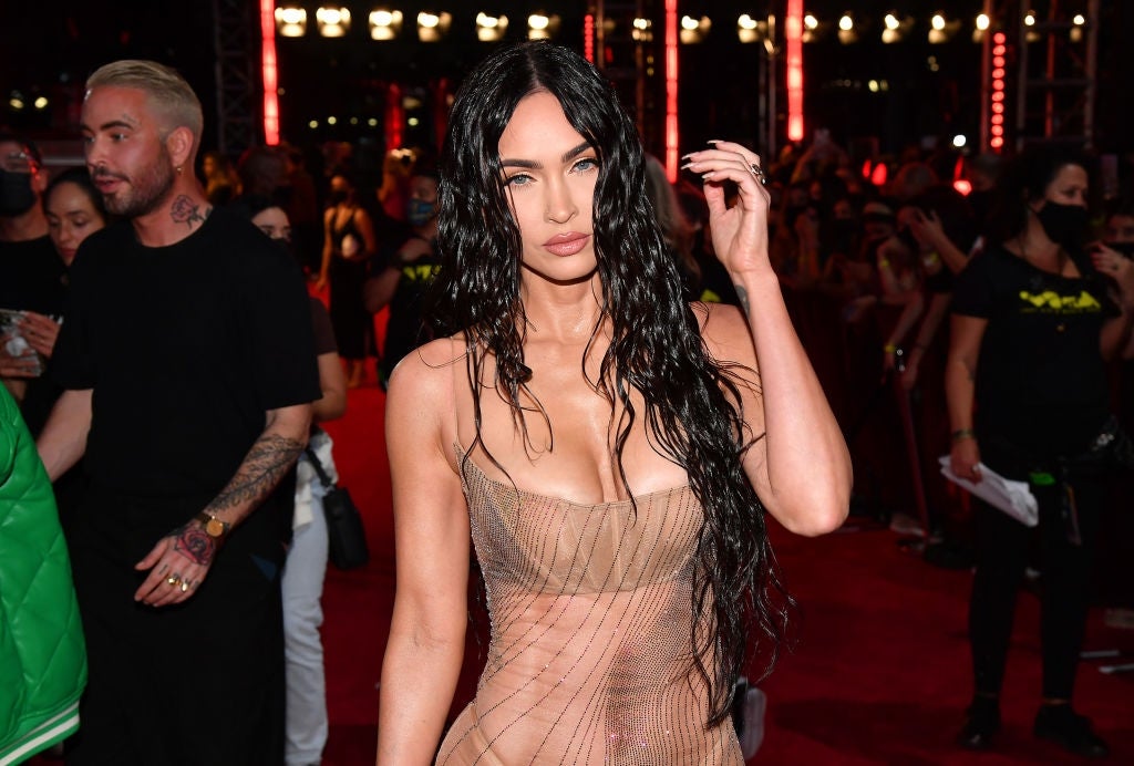 A person poses on the red carpet in a sheer, form-fitting dress with wet-look, wavy hair