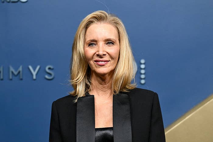 Person in elegant black satin-trimmed blazer stands against a backdrop with partial text "EMMYS."