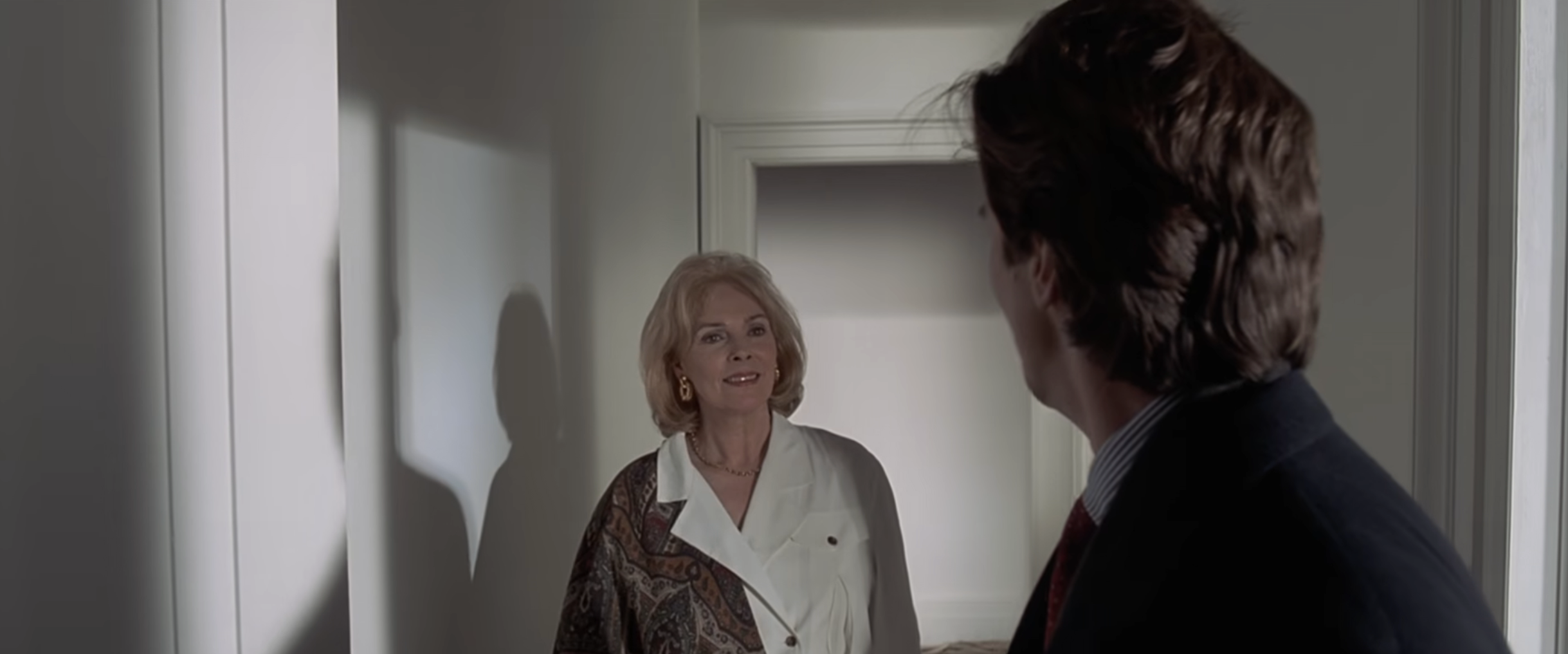 Misses Wolfe confronts Patrick Bateman, and she is wearing an elegant blouse, facing Bateman in a suit, standing in a hallway, in a scene from a movie