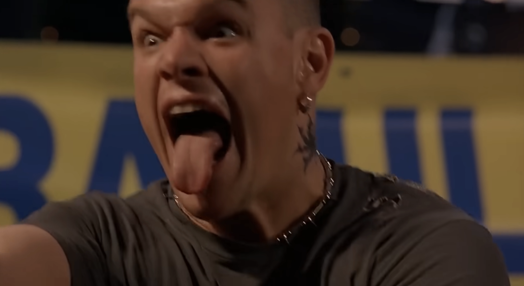 Person with a bold expression, sticking out their tongue, wearing a casual shirt and necklace, with a visible neck tattoo
