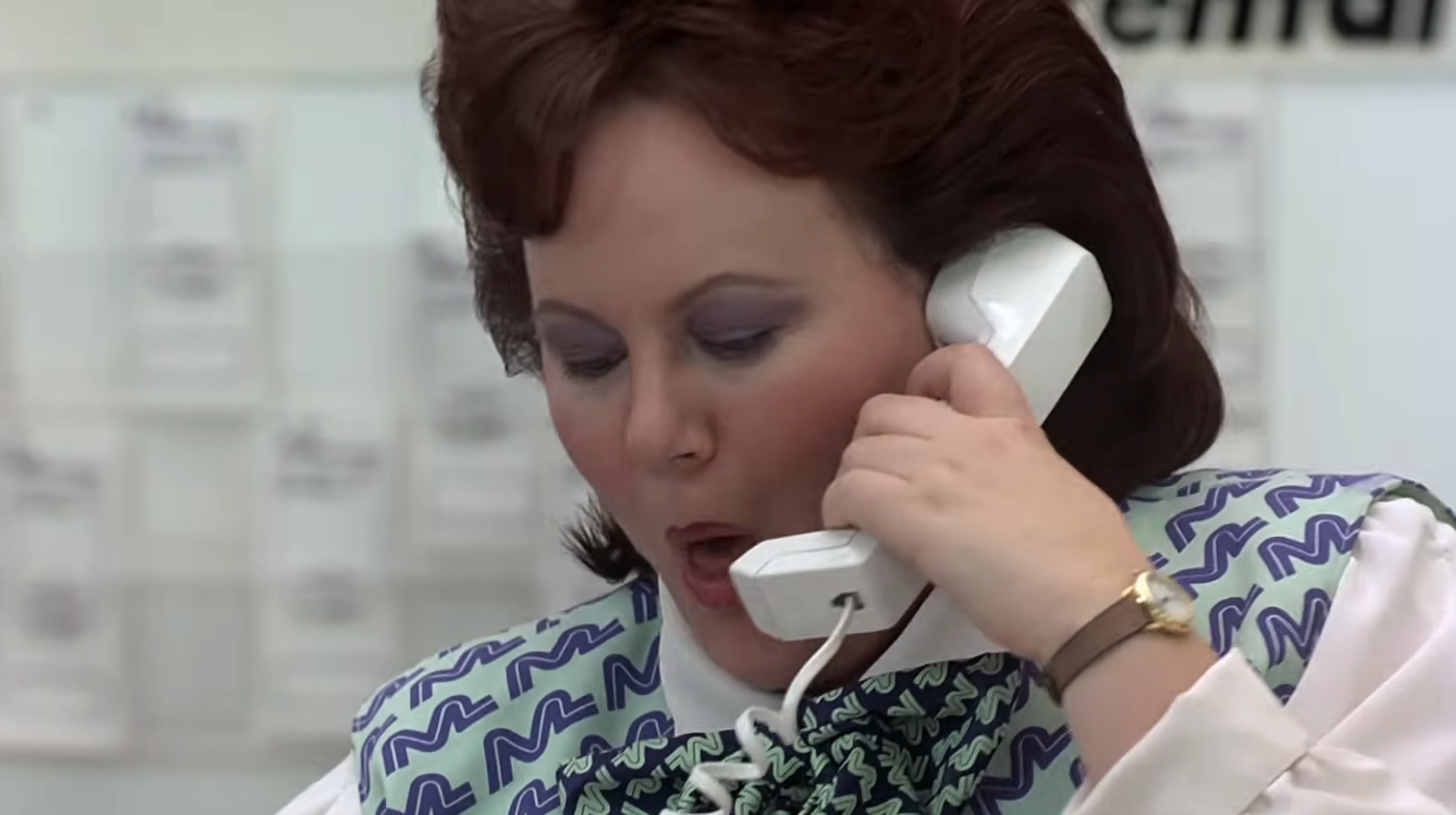 Person with a surprised expression talking on a corded phone, wearing a patterned vest over a white shirt