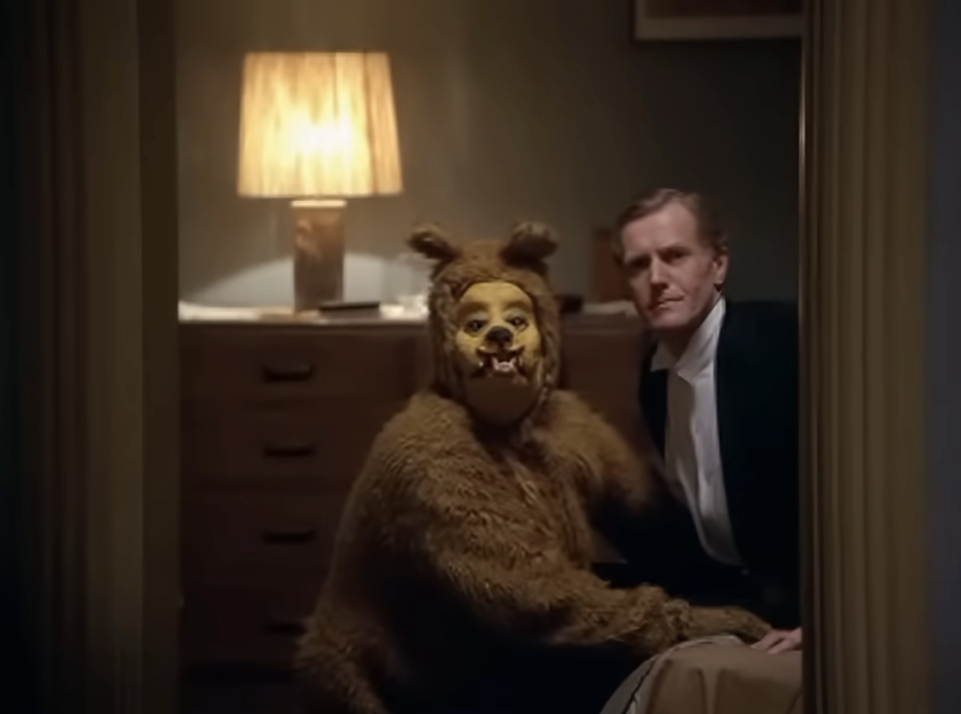 Person in bear costume crouches beside seated man in formal wear in dimly lit room. The scene suggests a whimsical or surreal setting