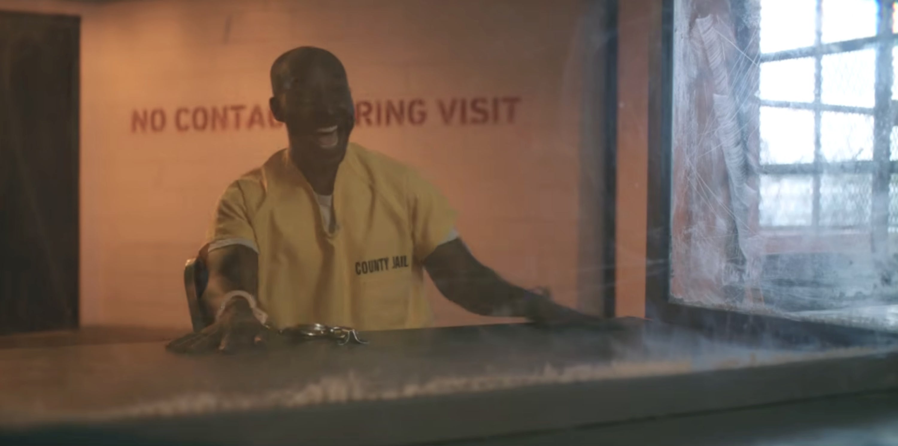 Man in yellow prison jumpsuit laughing, sitting at a visitation window with handcuffs on a table. Text reads "No Contact During Visit."