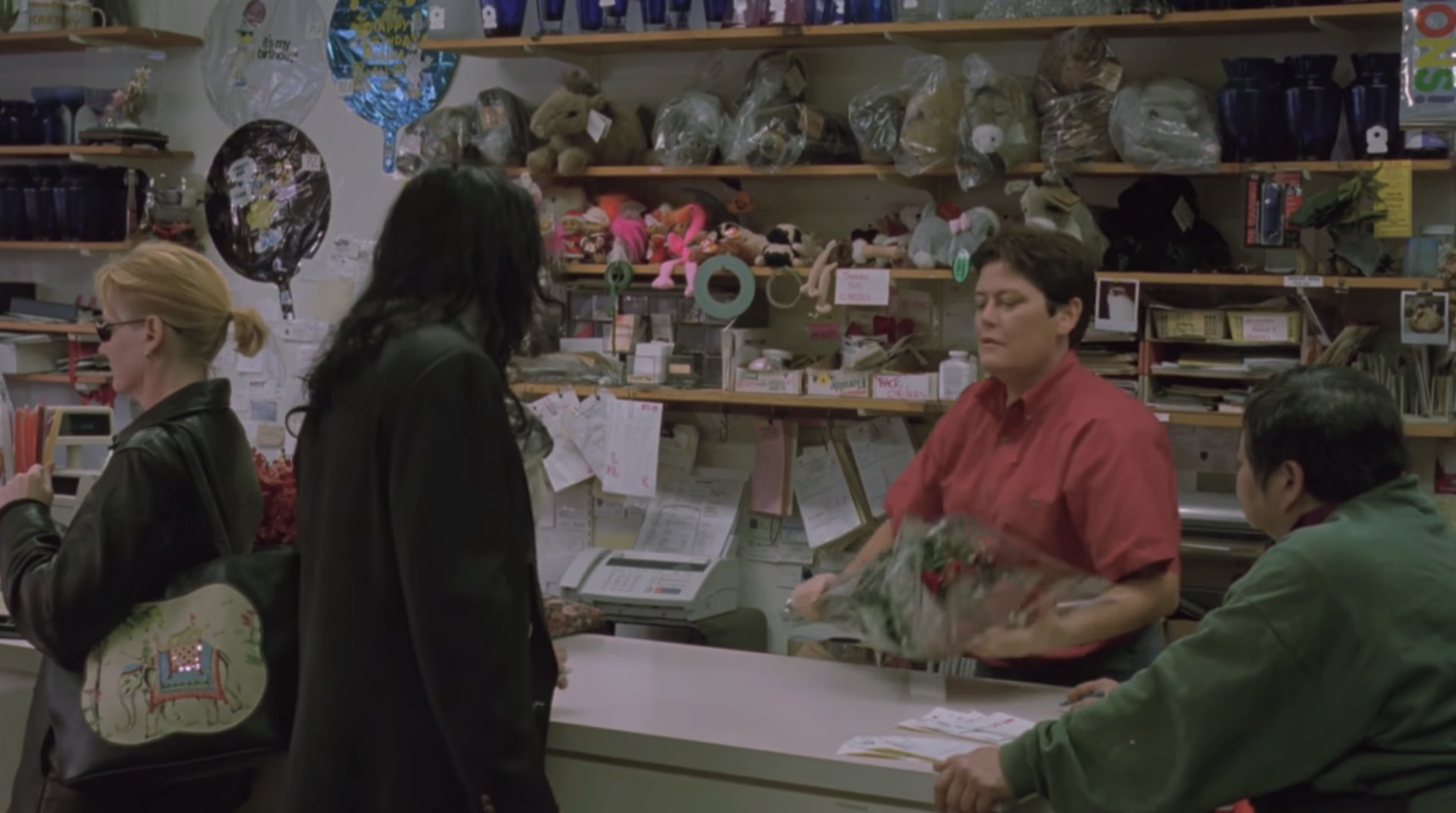 People in a flower shop, customer holding flowers at the counter amid shelves filled with stuffed animals and floral items