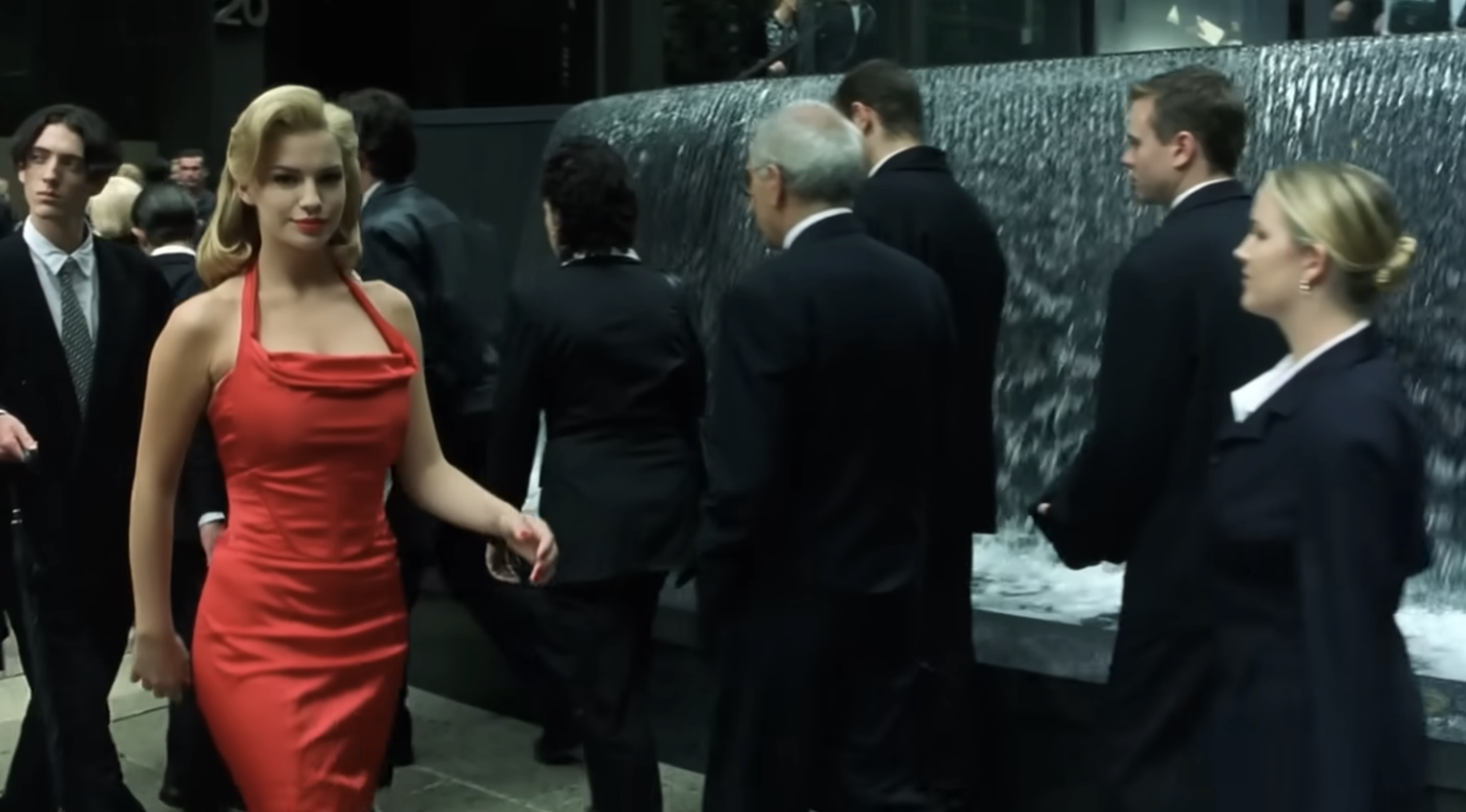 Person in a classic-style red dress walks past people in formal attire near a fountain