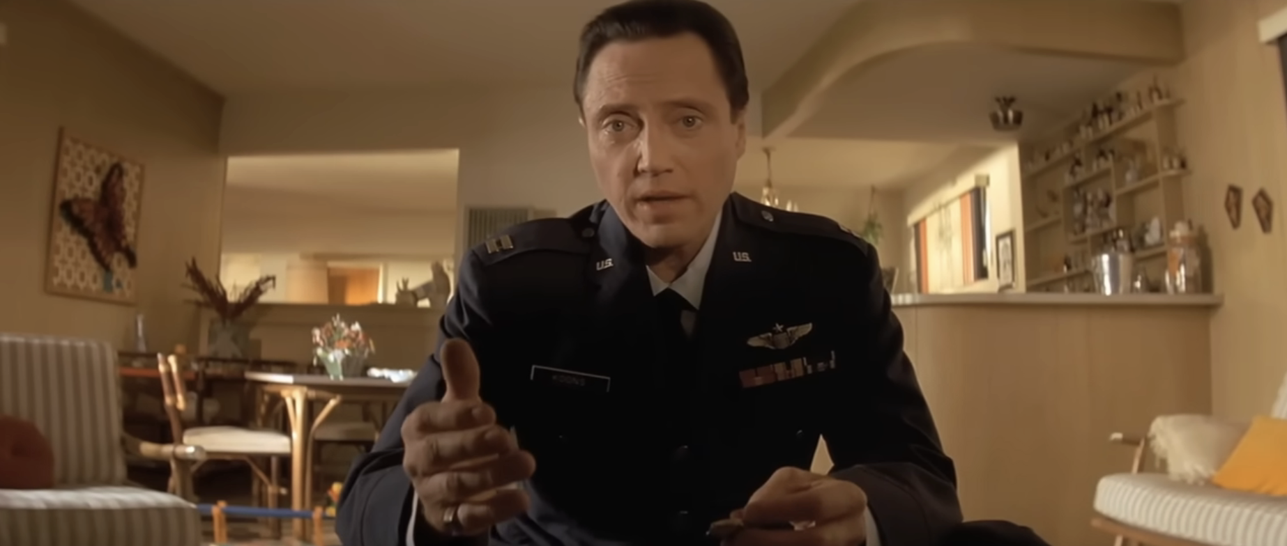 Person in military uniform sitting indoors, gesturing toward the camera. The setting appears to be a living room