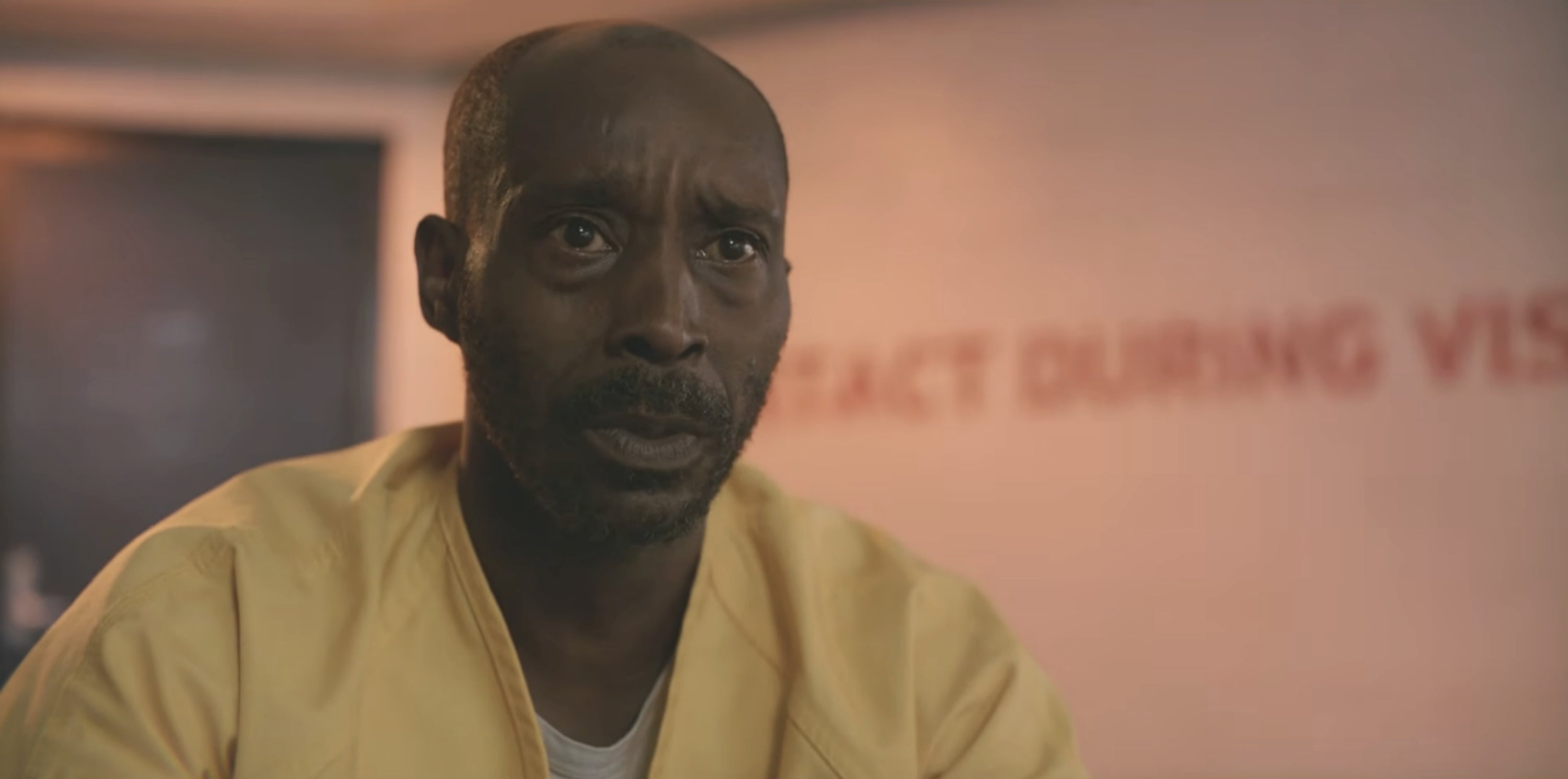 Man in a yellow prison uniform inside an interrogation room, looking seriously at someone off-camera