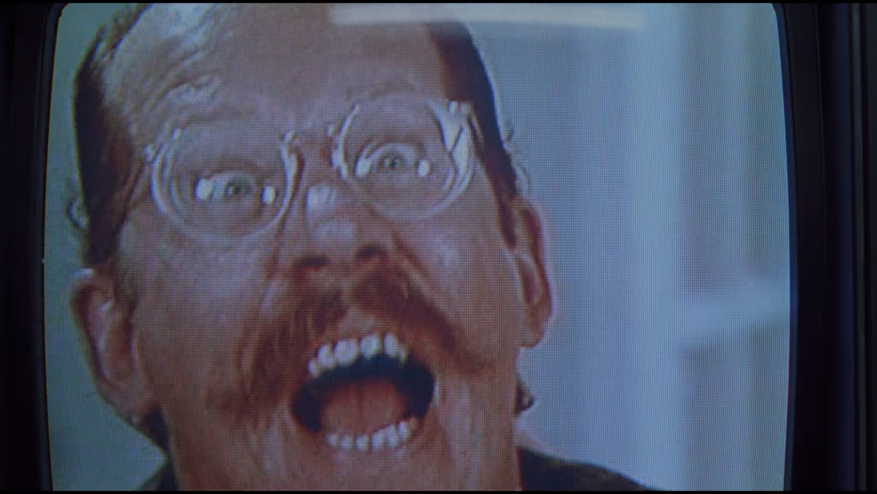 A man with glasses and a mustache appears on a TV screen, looking amazed with mouth open wide