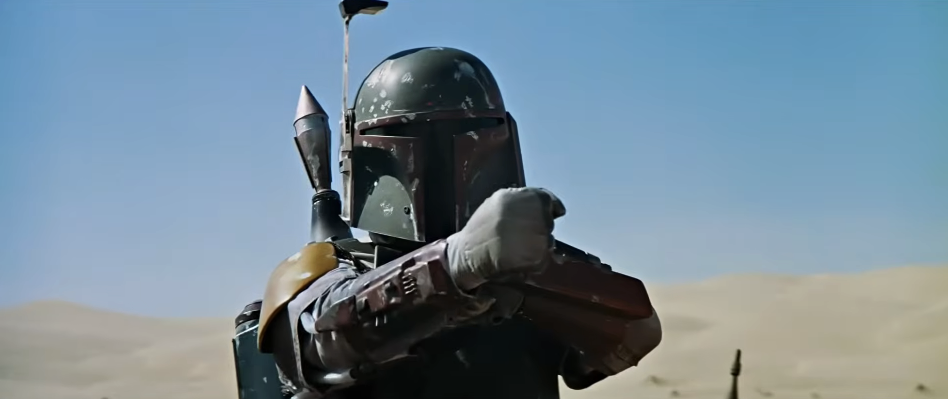A person dressed in futuristic armor stands in a desert setting, pointing forward, resembling a character from a sci-fi movie