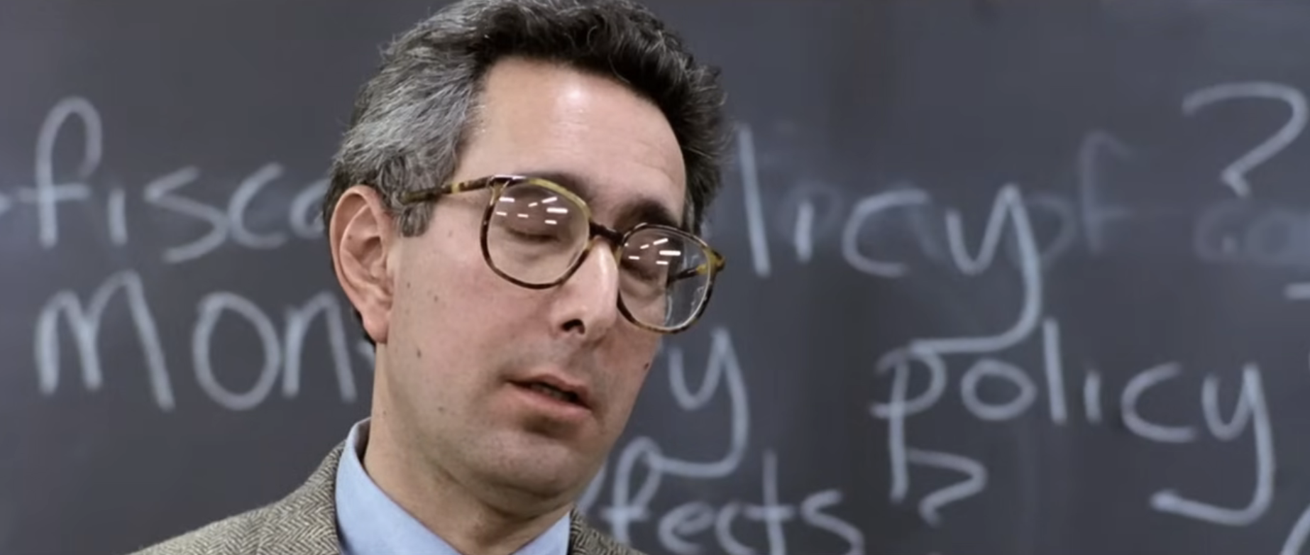A man with glasses and a bemused expression stands in front of a chalkboard with economics-related terms