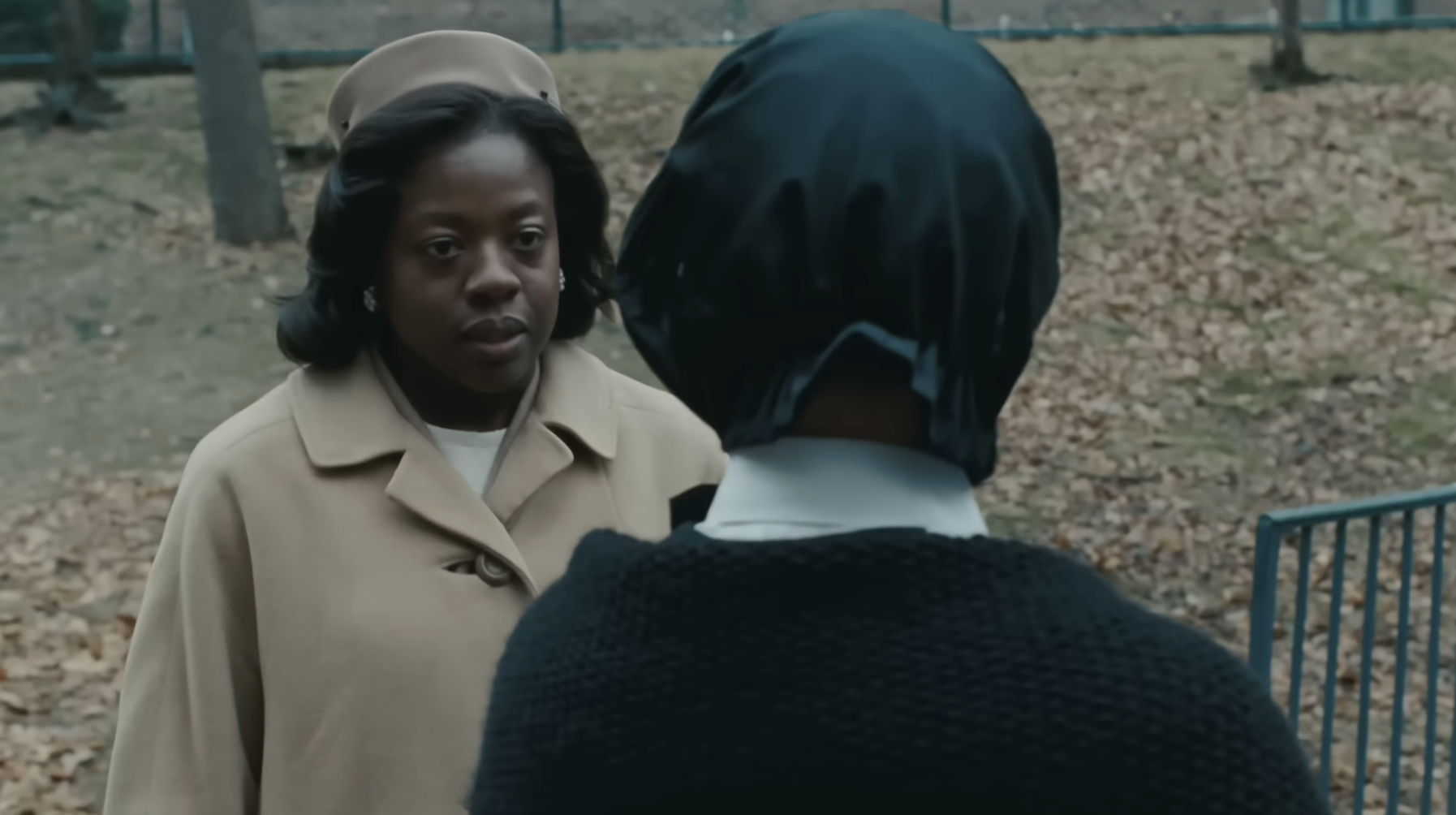 Two characters in a park have a serious conversation. One wears a coat and hat, the other a headscarf and knit top. The scene is tense and dramatic