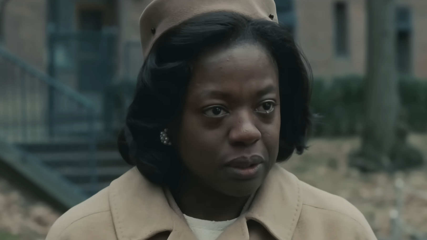 Woman in a serious expression wearing a vintage-style coat and hat, standing outdoors