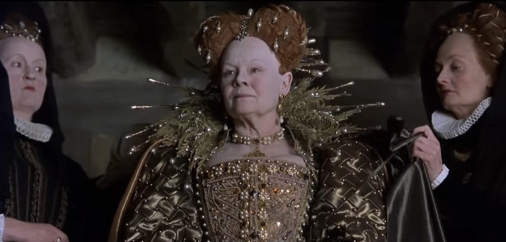 Three actors in Elizabethan costumes, with intricate headpieces and ornate gowns, in a historical film scene
