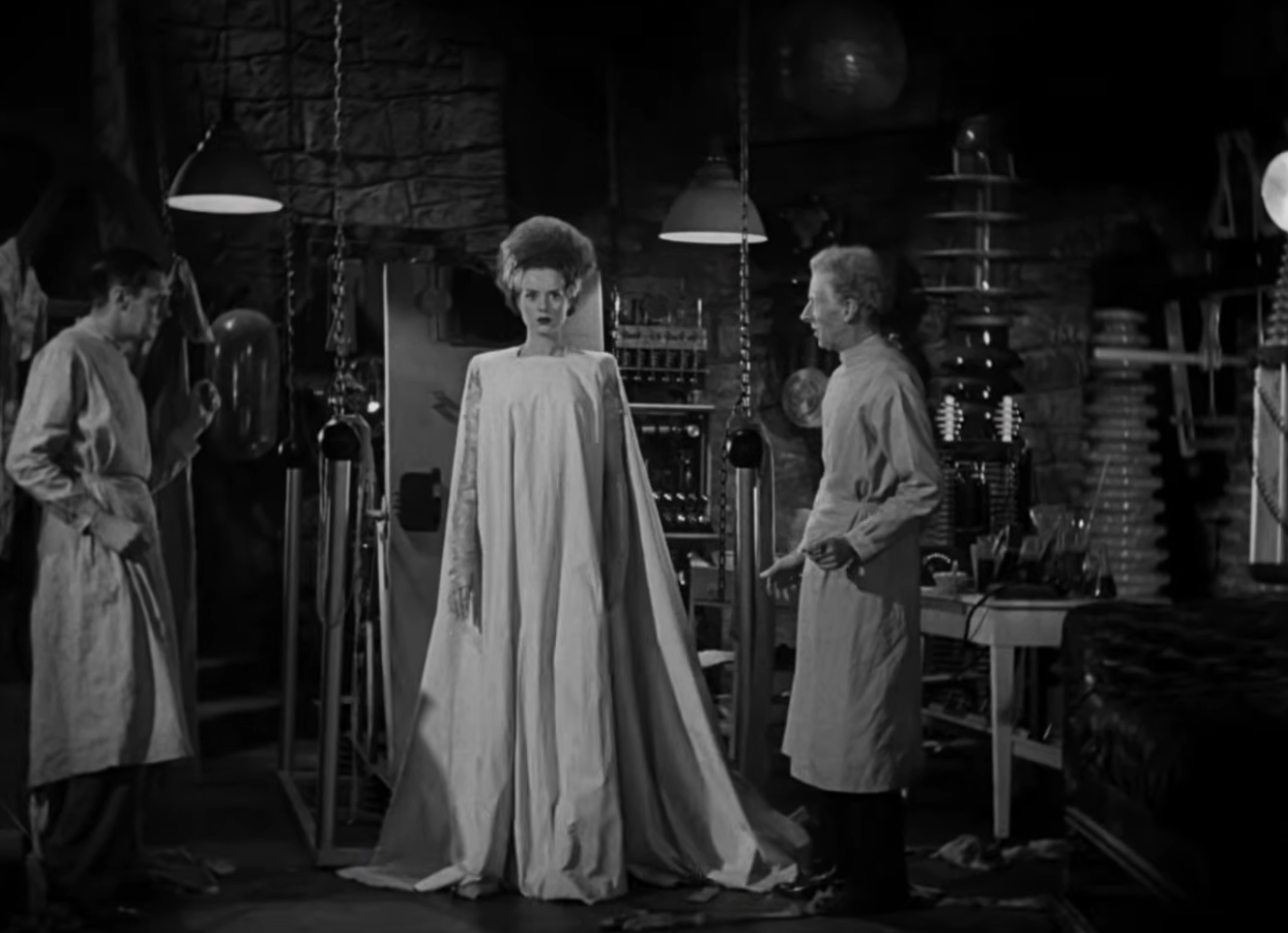 A classic movie scene depicting a bride-like figure in a long, flowing gown standing in a laboratory with two men in lab coats