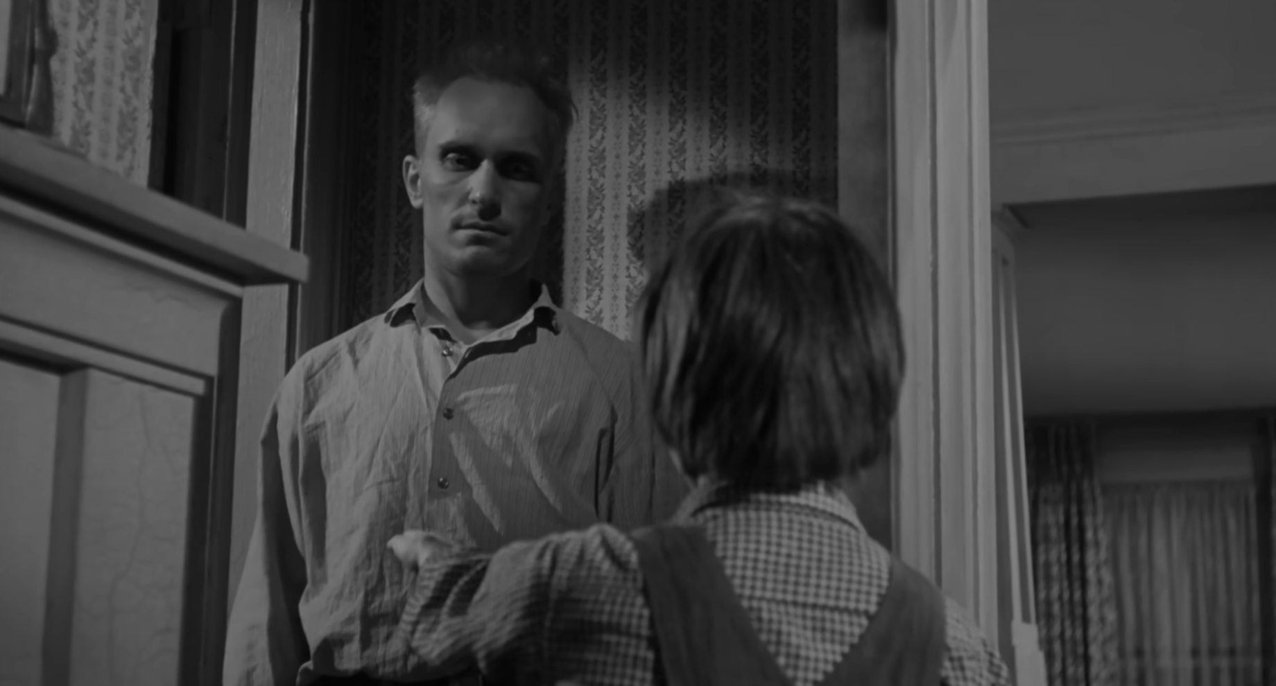 A man stands silently facing a child in a tense moment. The background features patterned wallpaper and curtains