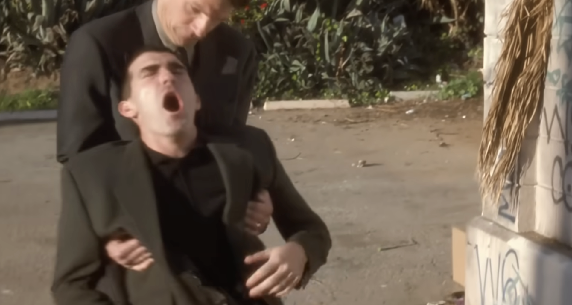 Person in formal attire appears distressed while being held by another person outdoors