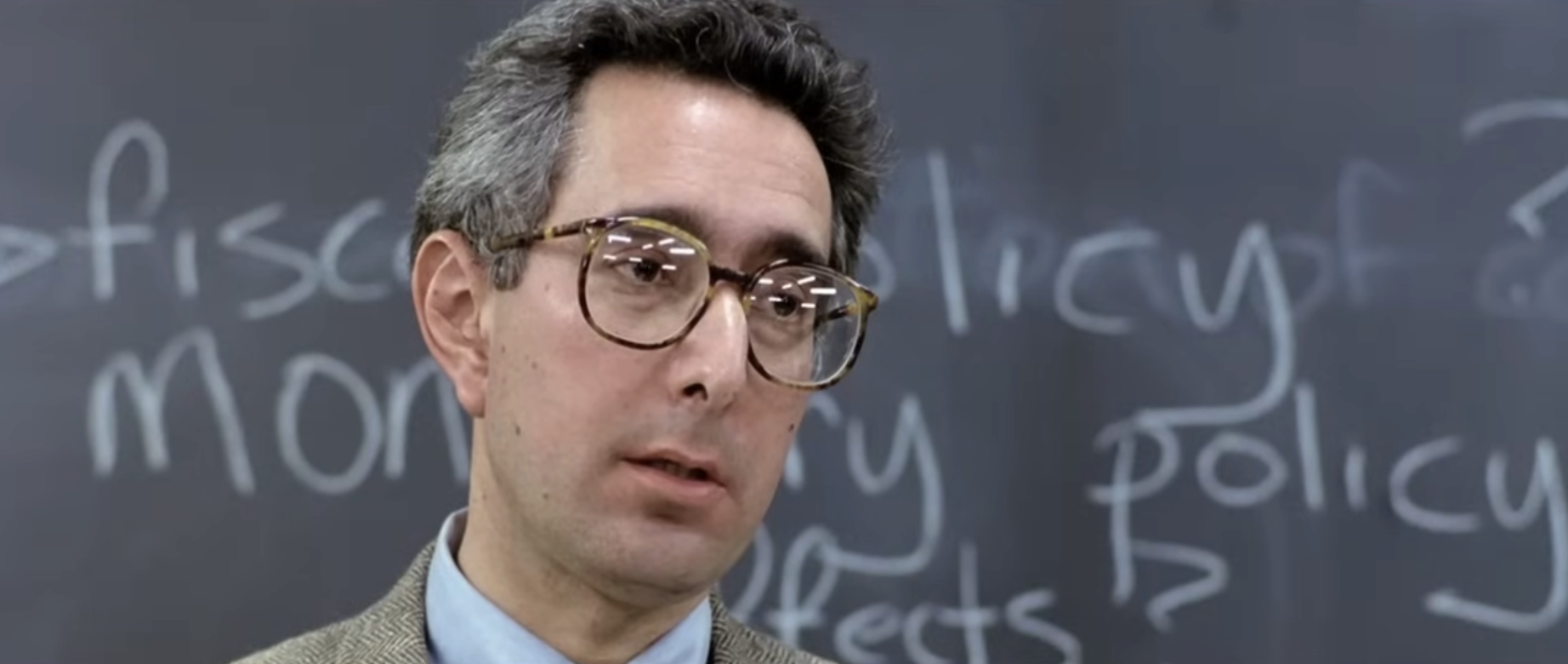 A man in glasses speaks in front of a chalkboard with semi-visible writing