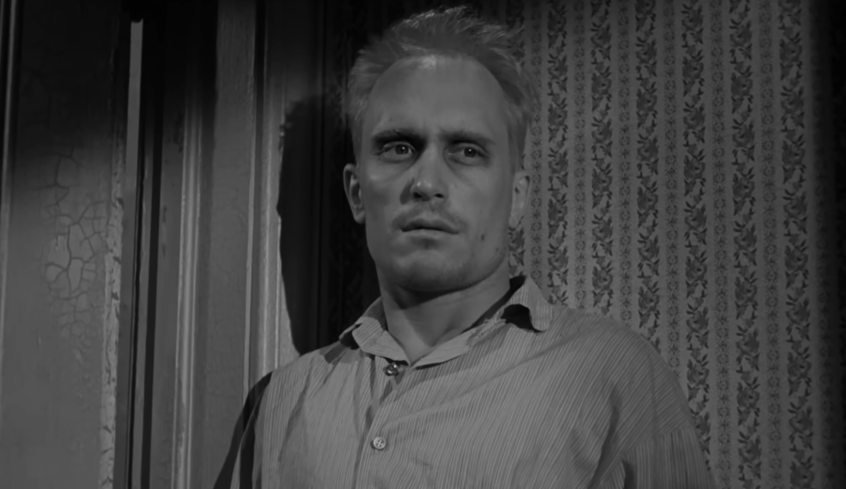 Scene from a classic black-and-white film featuring a man with short hair and a serious expression standing against a wallpapered wall