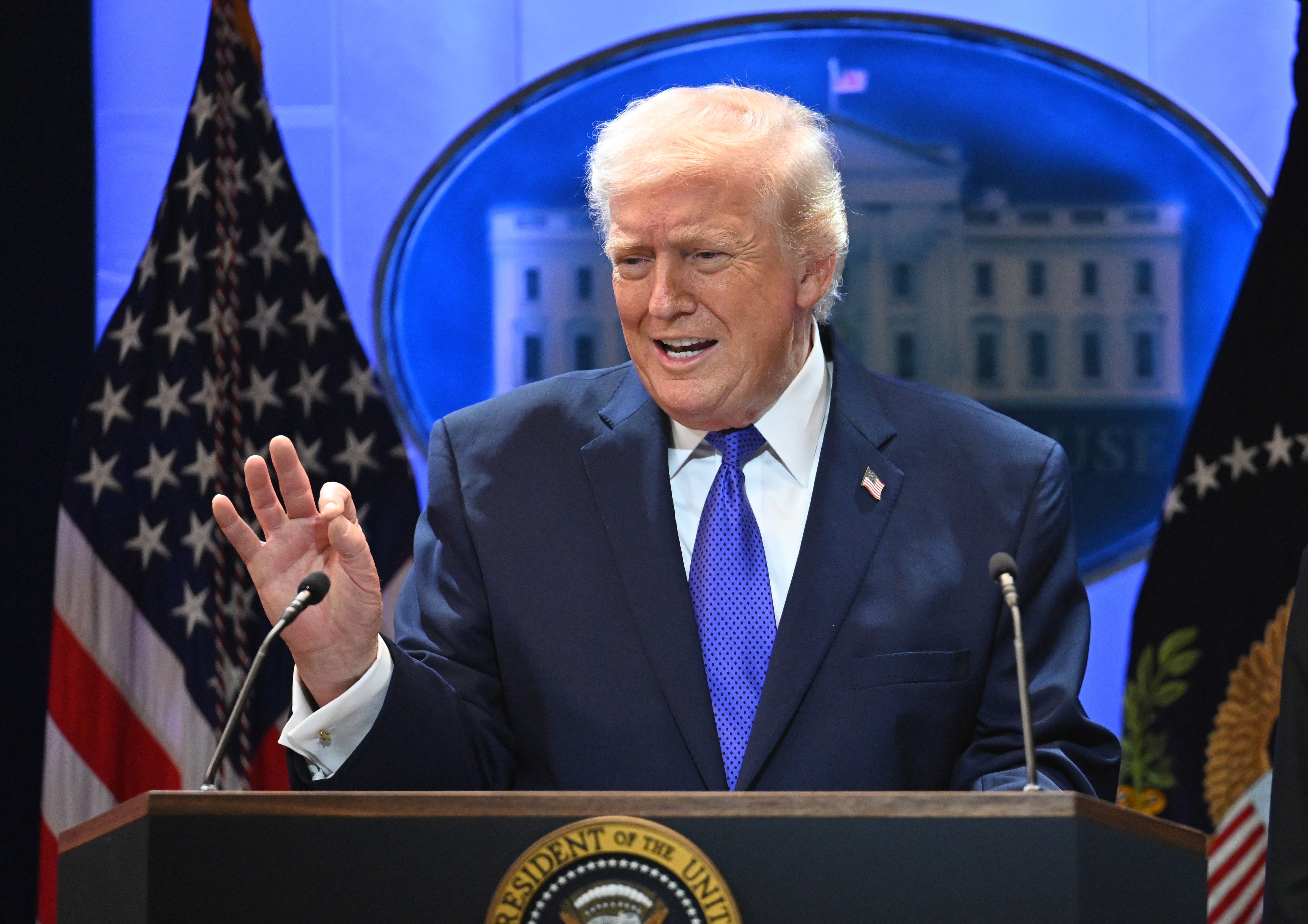 En person i dress taler ved et podium med den amerikanske presidentens segl, flankert av flagg og et bilde av Det hvite hus i bakgrunnen