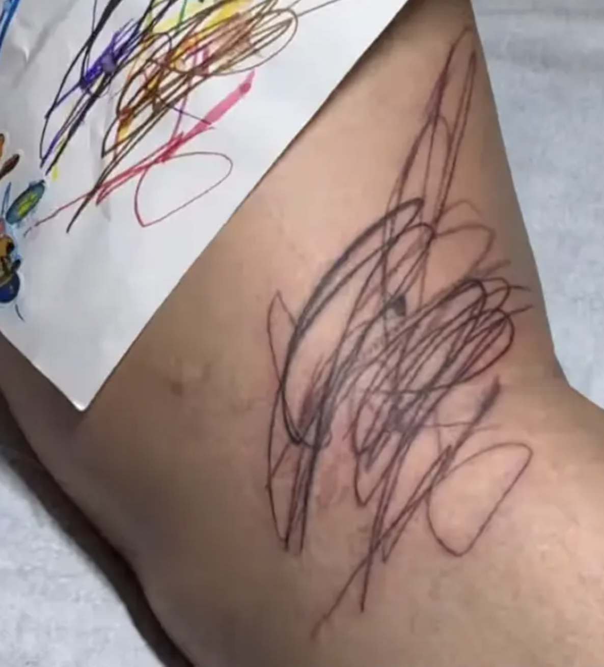 Child's colorful scribble on paper; tattooed scribble on skin mimics it. Artistic expression theme