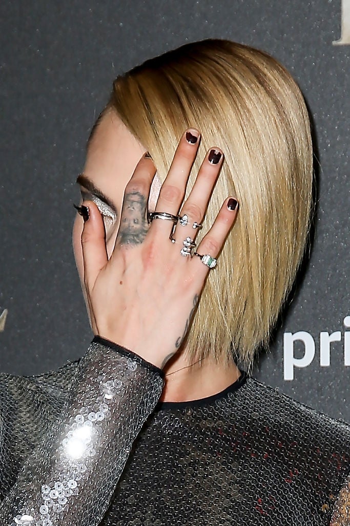 Person with short hair covers face with hand, showing multiple rings and tattoos. Wears a sequined outfit at a celebrity event