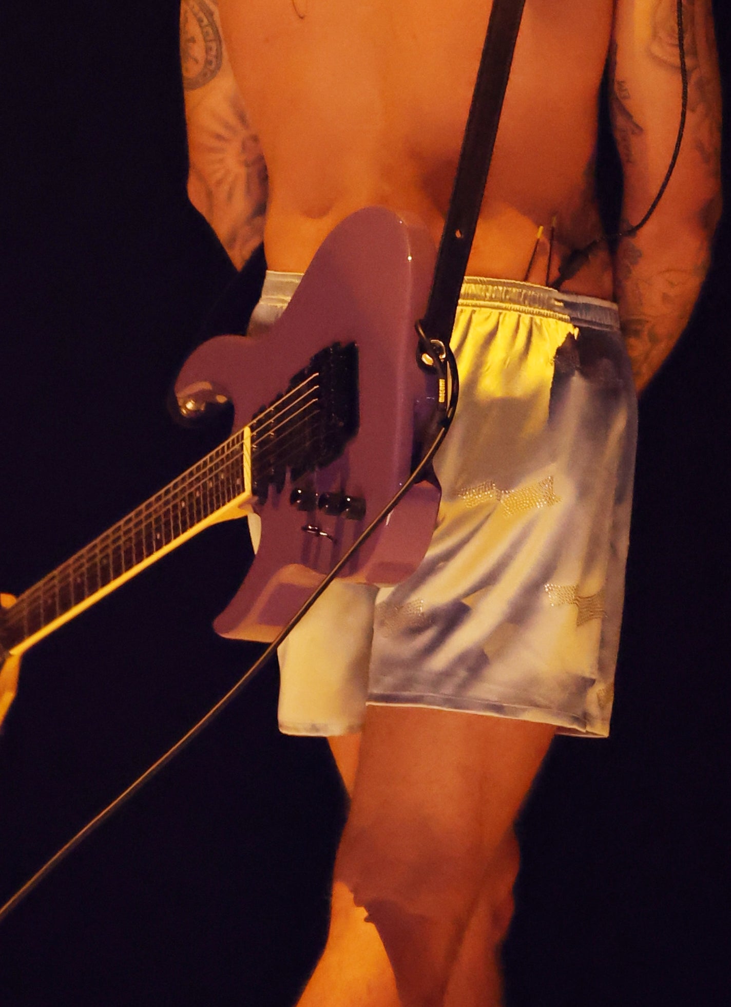 Musician on stage wearing shorts, shirtless, with tattoos visible and a guitar on their back, creating a casual, rock-inspired look