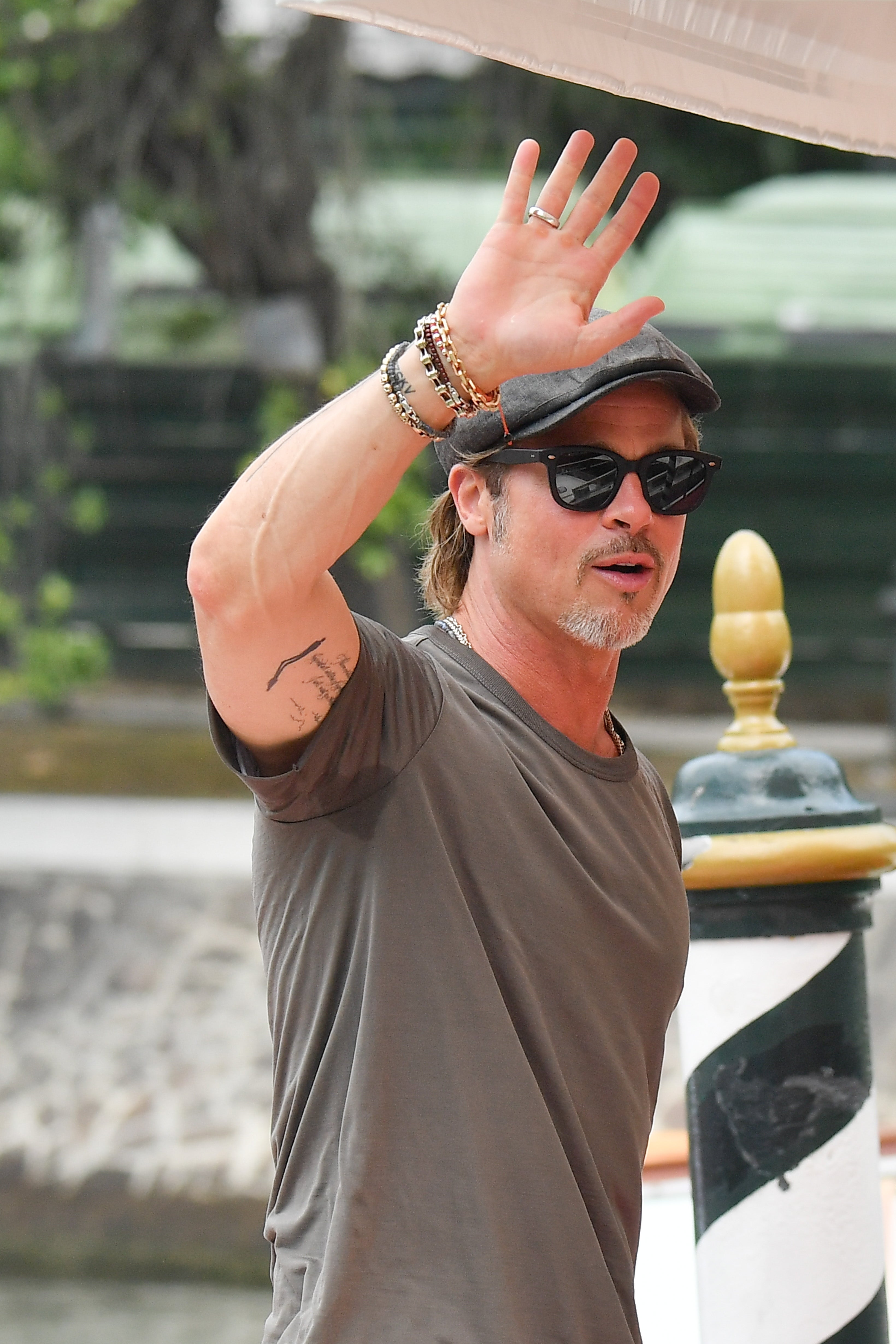 A person wearing sunglasses, a flat cap, a T-shirt, and bracelets is waving outdoors