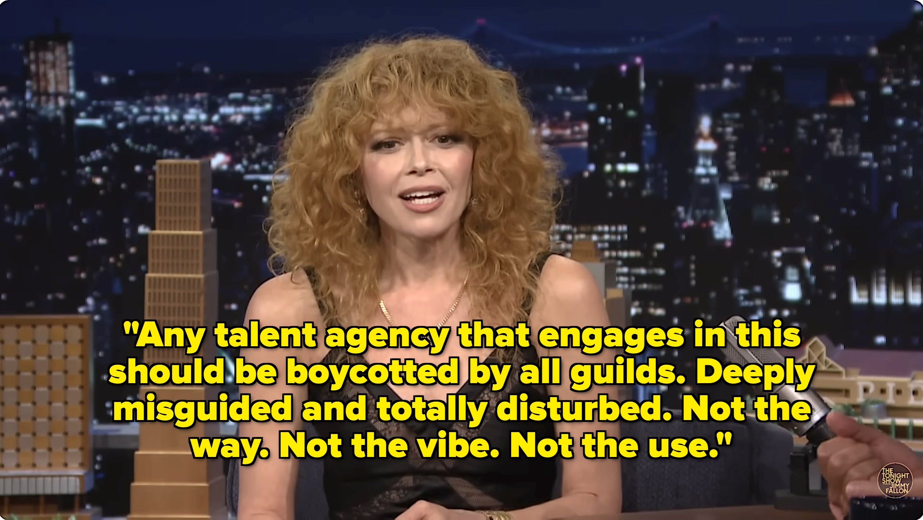 Person with curly hair speaking on a talk show, wearing a sleeveless top with lace detail. Microphone visible