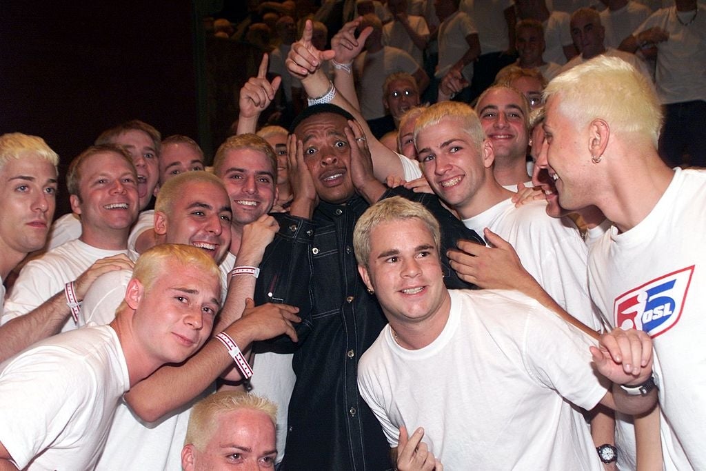 Group of fans with blond hair surround a person, who looks surprised or entertained, wearing a black outfit