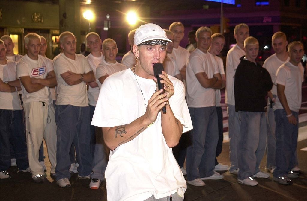 A person in a white shirt and cap speaks into a microphone, surrounded by a group wearing similar outfits
