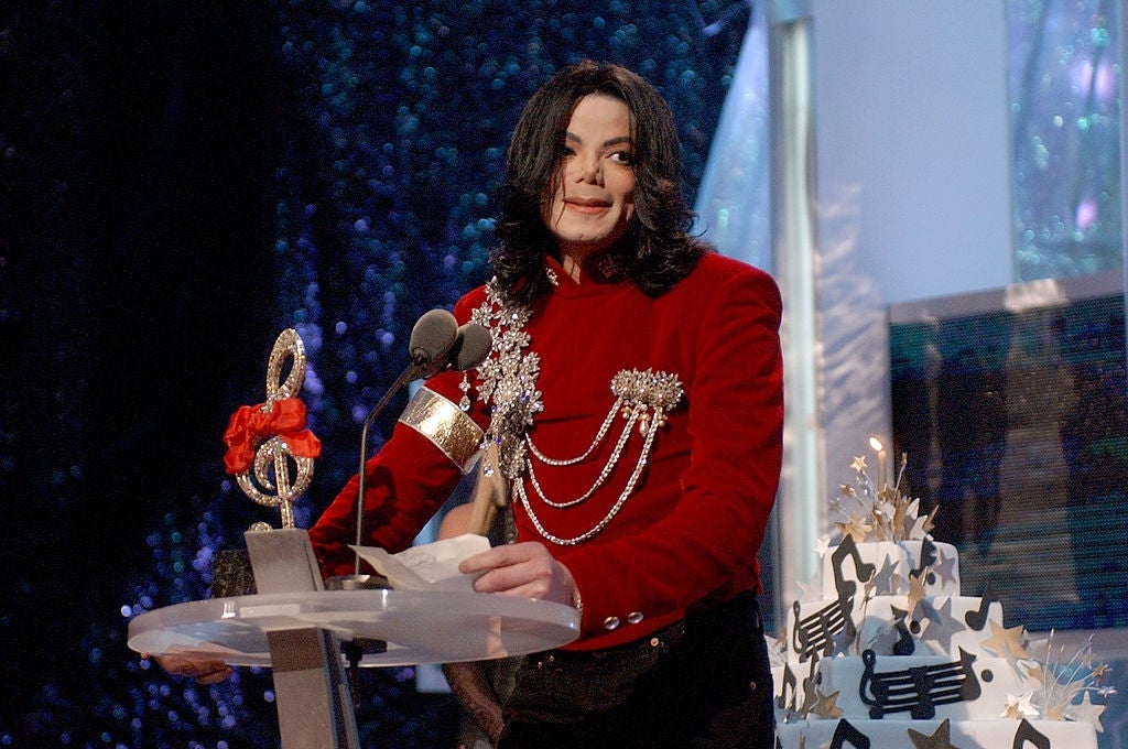 Person in ornate red outfit with embellishments speaks at a podium, a shiny background and a cake with musical notes in the foreground