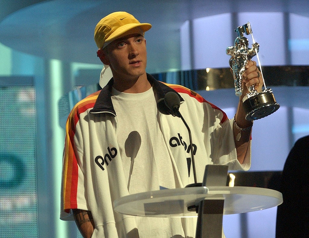 Person holding an award, wearing a casual cap and oversized sports jacket, speaking at a podium during an event